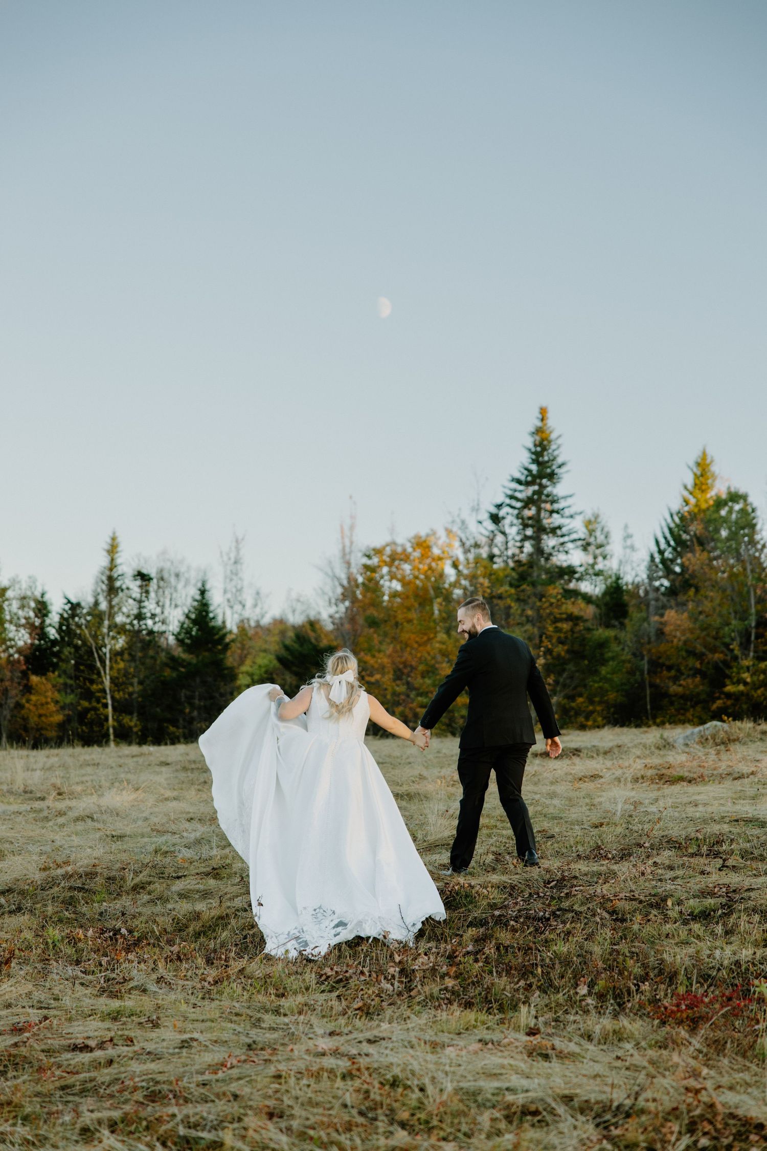 Wedding couple walks through an autumn field with trees and blue sky in the background.