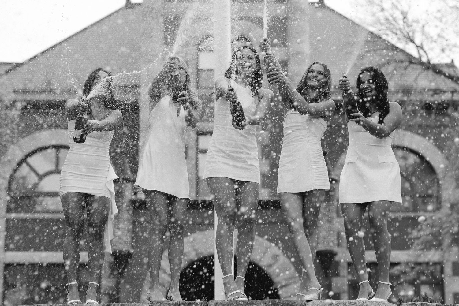 Five women in white dresses celebrate outdoors with confetti flying around them in a black and white photo.