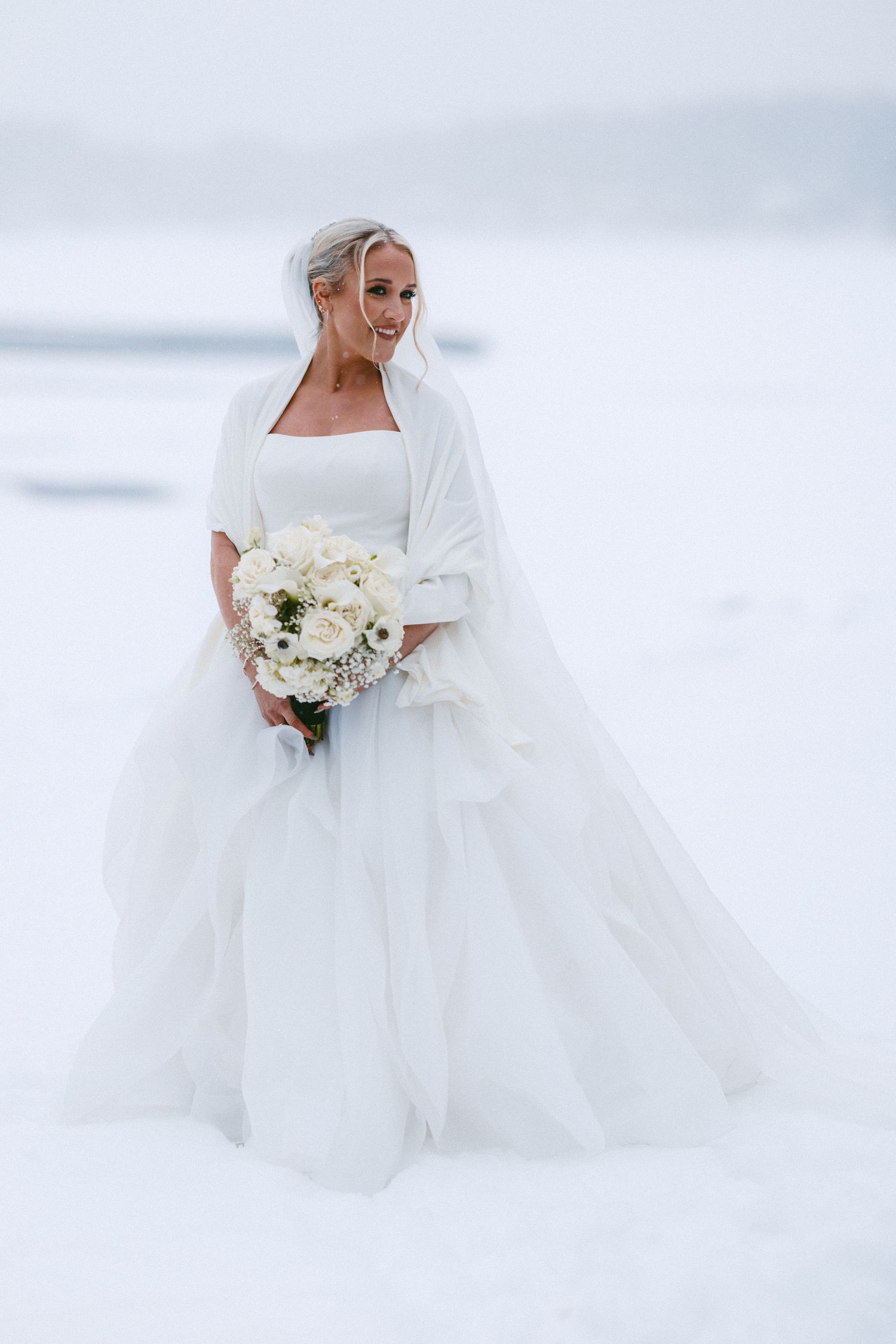 Ethereal winter bridal portraits show a flowing white wedding dress and bouquet against a snowy landscape.
