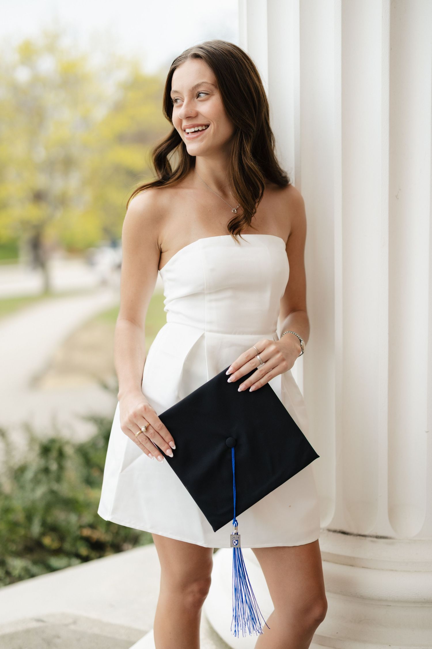 Graduate posing with decorated graduation cap and white dress against white columns.