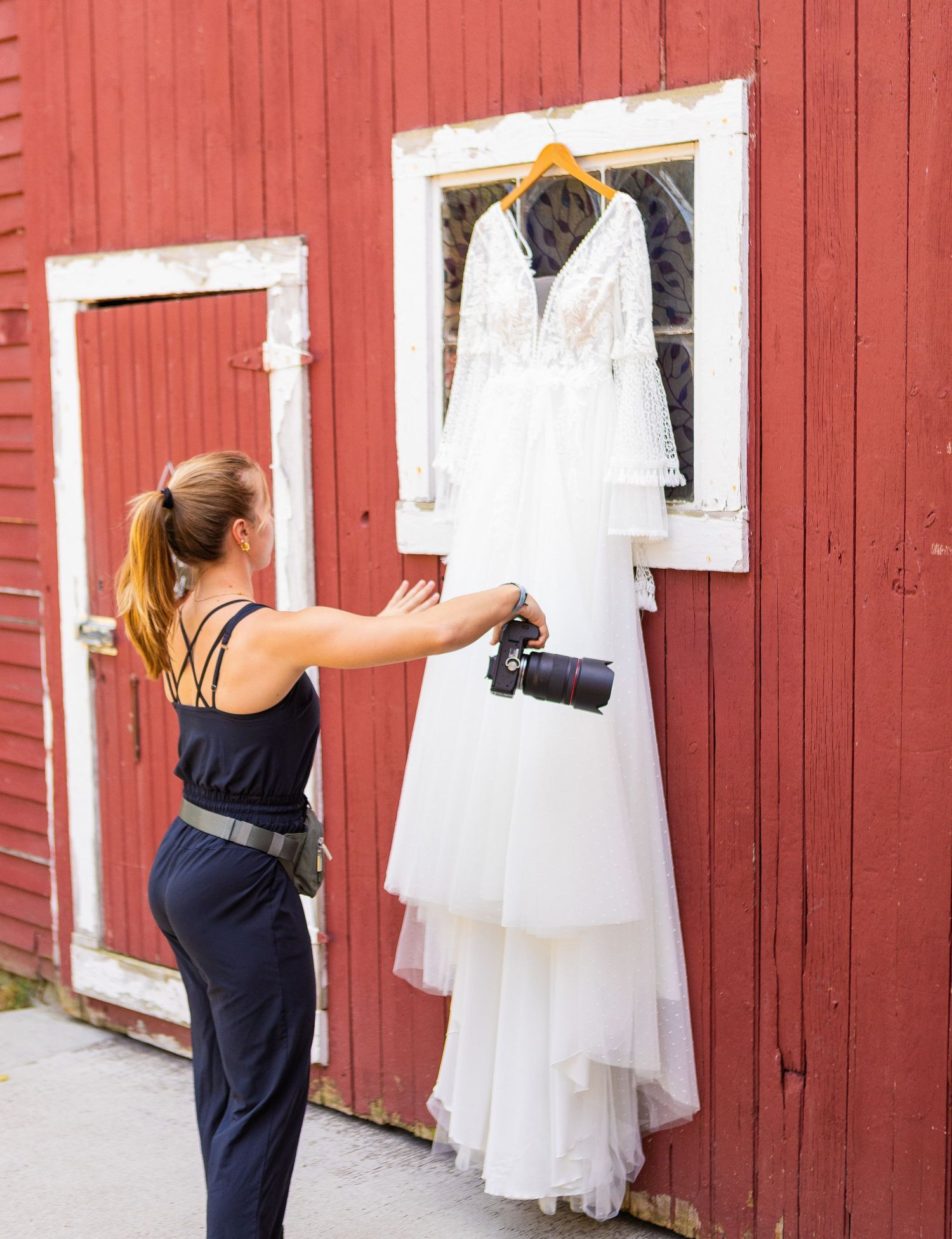 A photographer in a navy dress takes pictures of white wedding gowns hanging on a red barn wall.