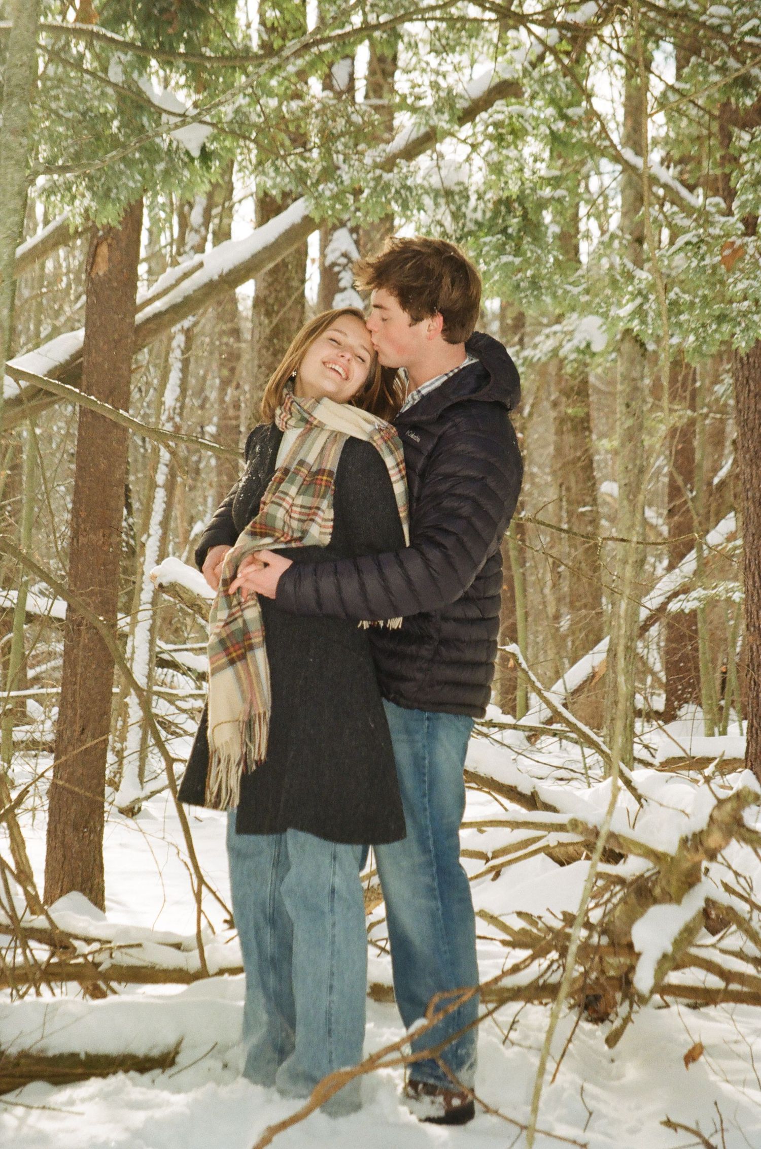 A couple shares romantic moments in a snowy forest setting with pine trees in winter.