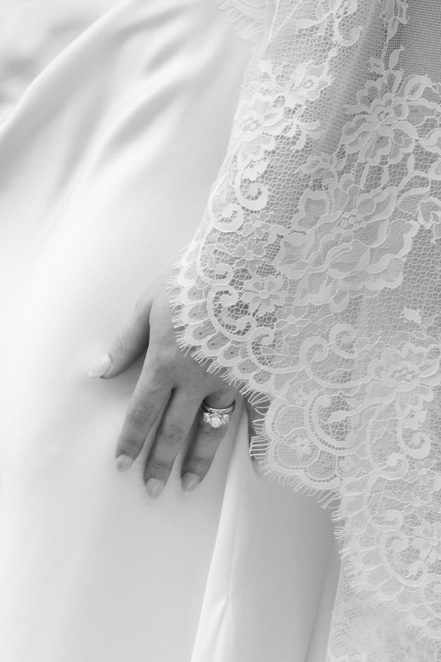 Close-up of bride's hand with diamond engagement ring on white lace wedding dress in black and white.
