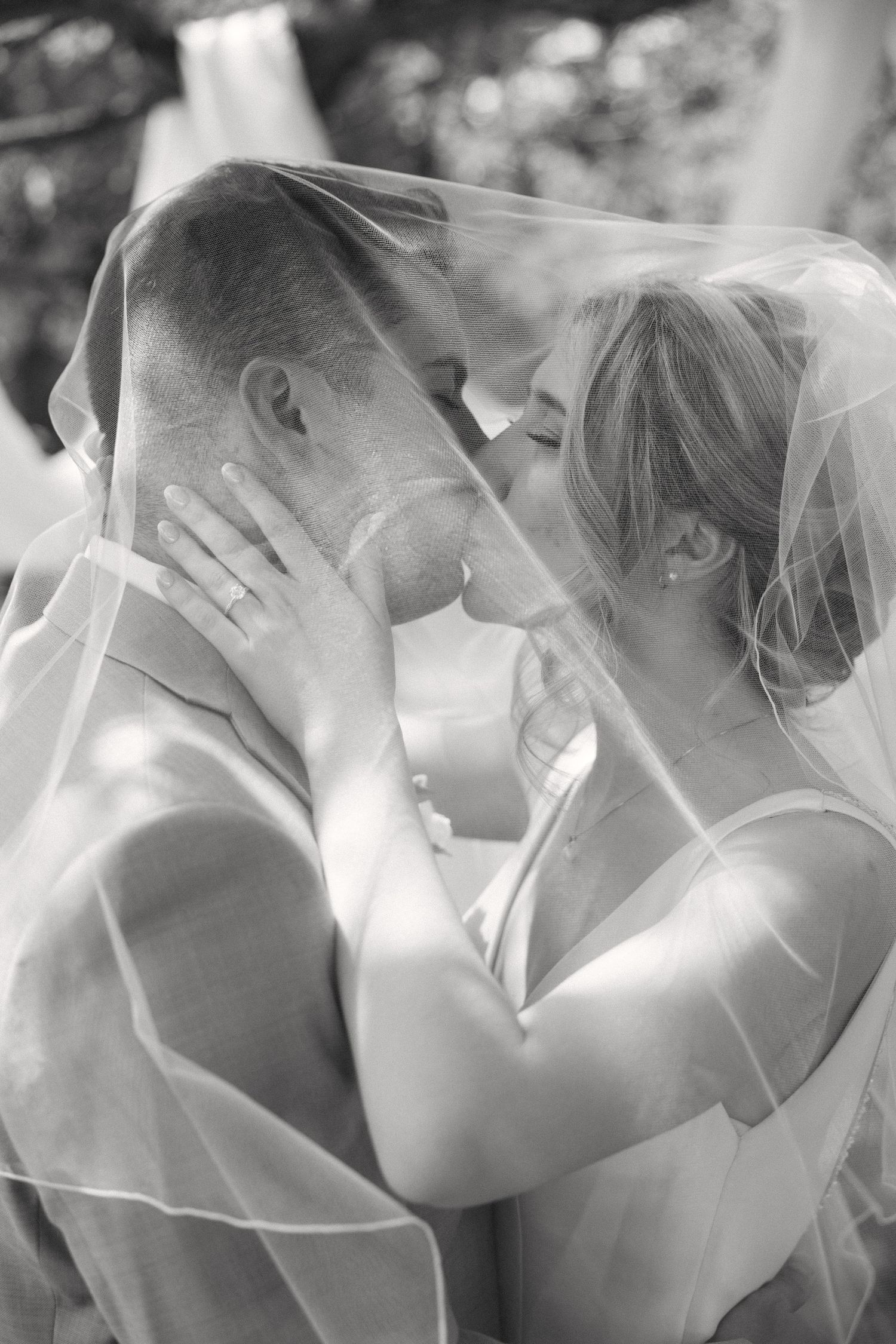 Black and white photo of bride and groom sharing a kiss under a wedding veil outdoors.