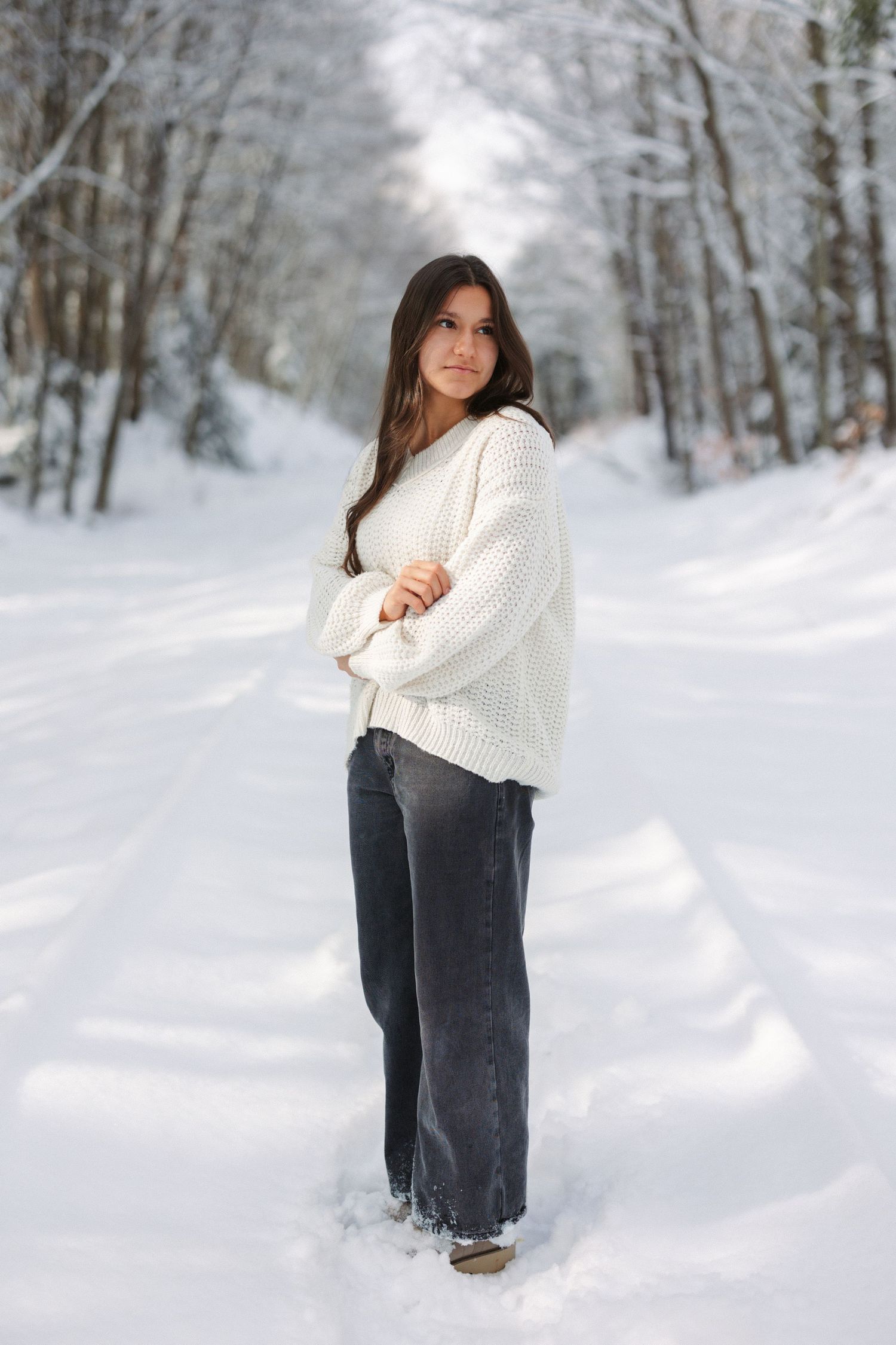 A person in a cozy white sweater and dark pants stands on a snow-covered path surrounded by winter trees.