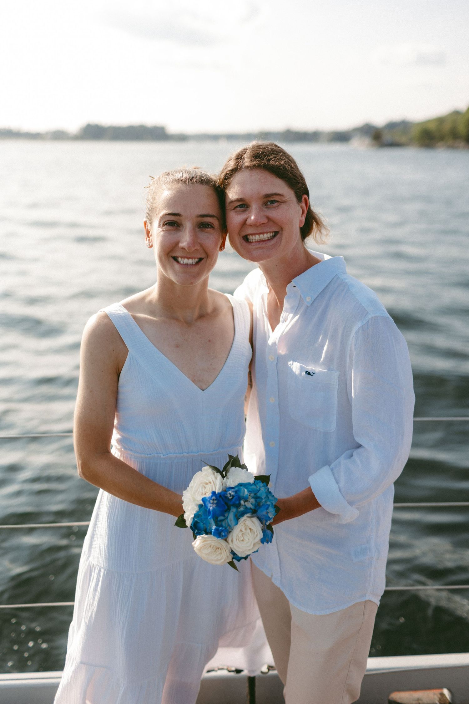 A couple in matching white attire holds a blue and white bouquet by the water.