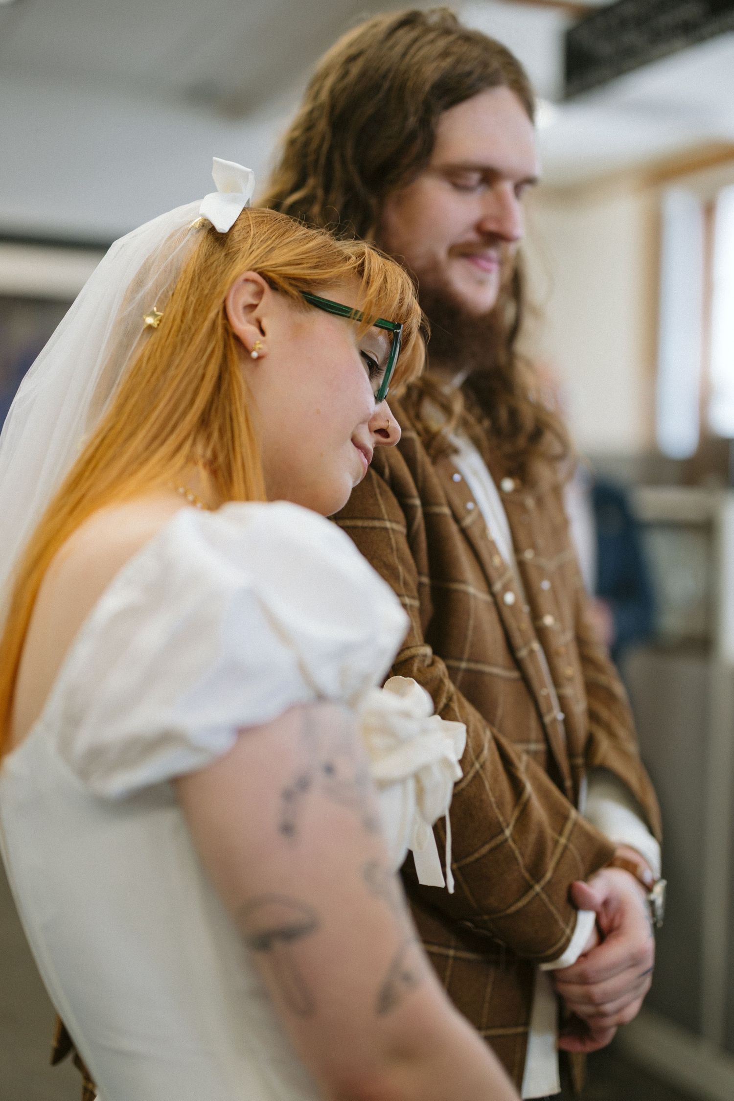 A tattooed bride in white dress and veil with green glasses stands beside a groom in brown plaid suit at a wedding ceremony.