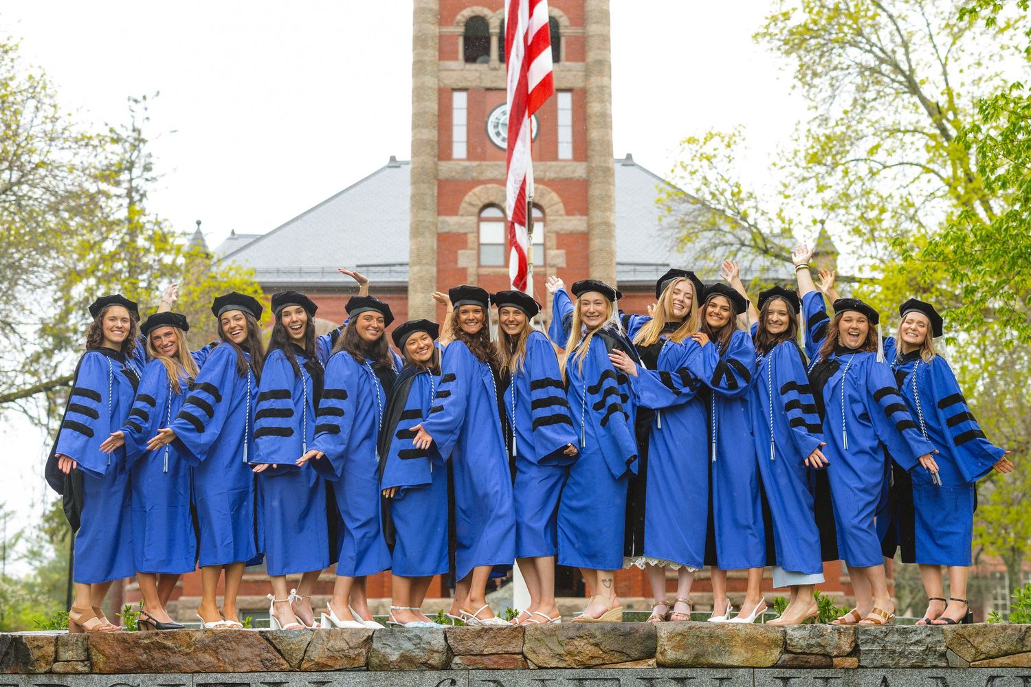 Group of graduates in blue caps and gowns celebrating outdoors near a brick bell tower on graduation day.