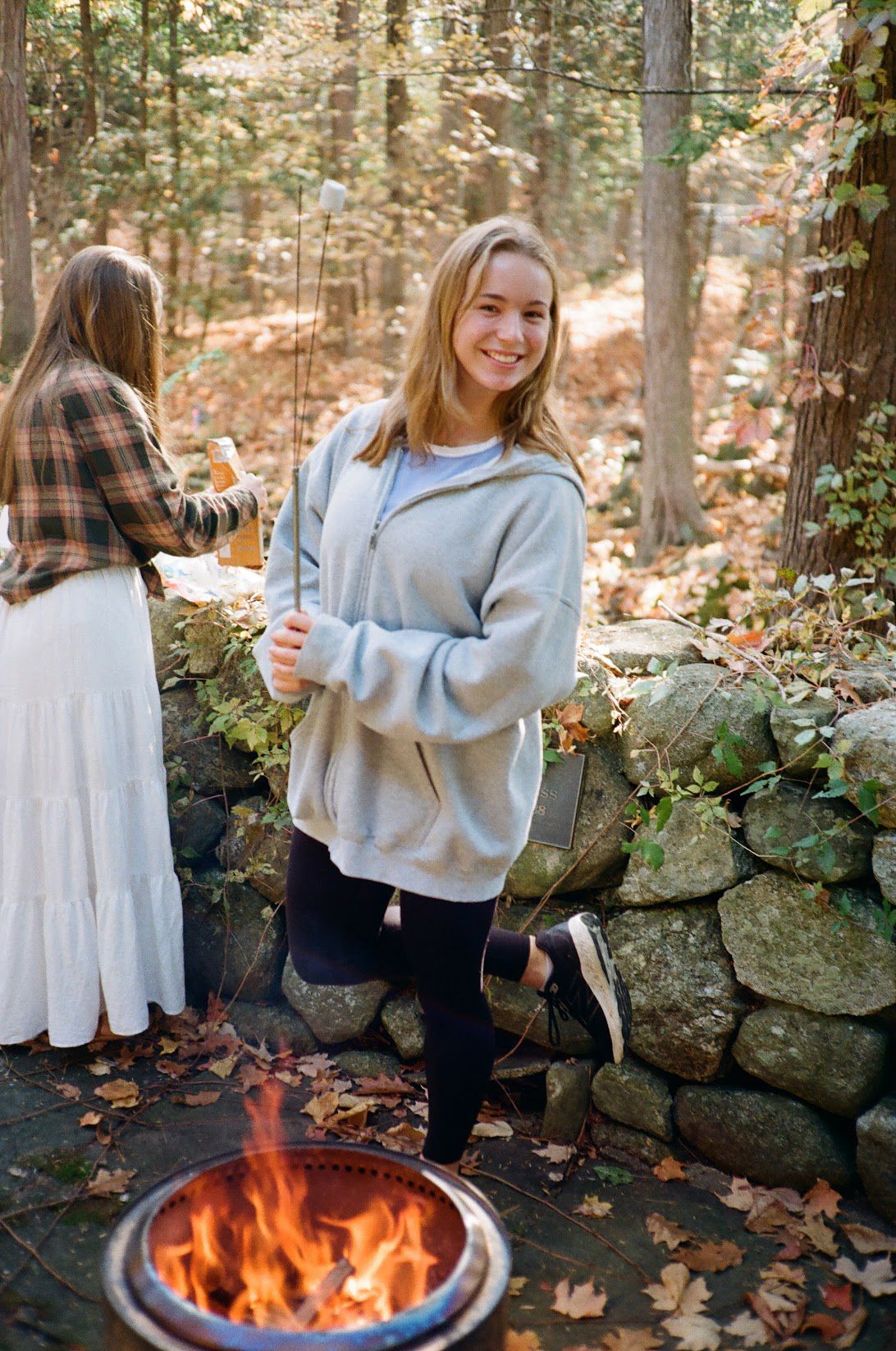 Person in cozy sweater enjoying outdoor fall activities by stone wall and fire pit in wooded setting.
