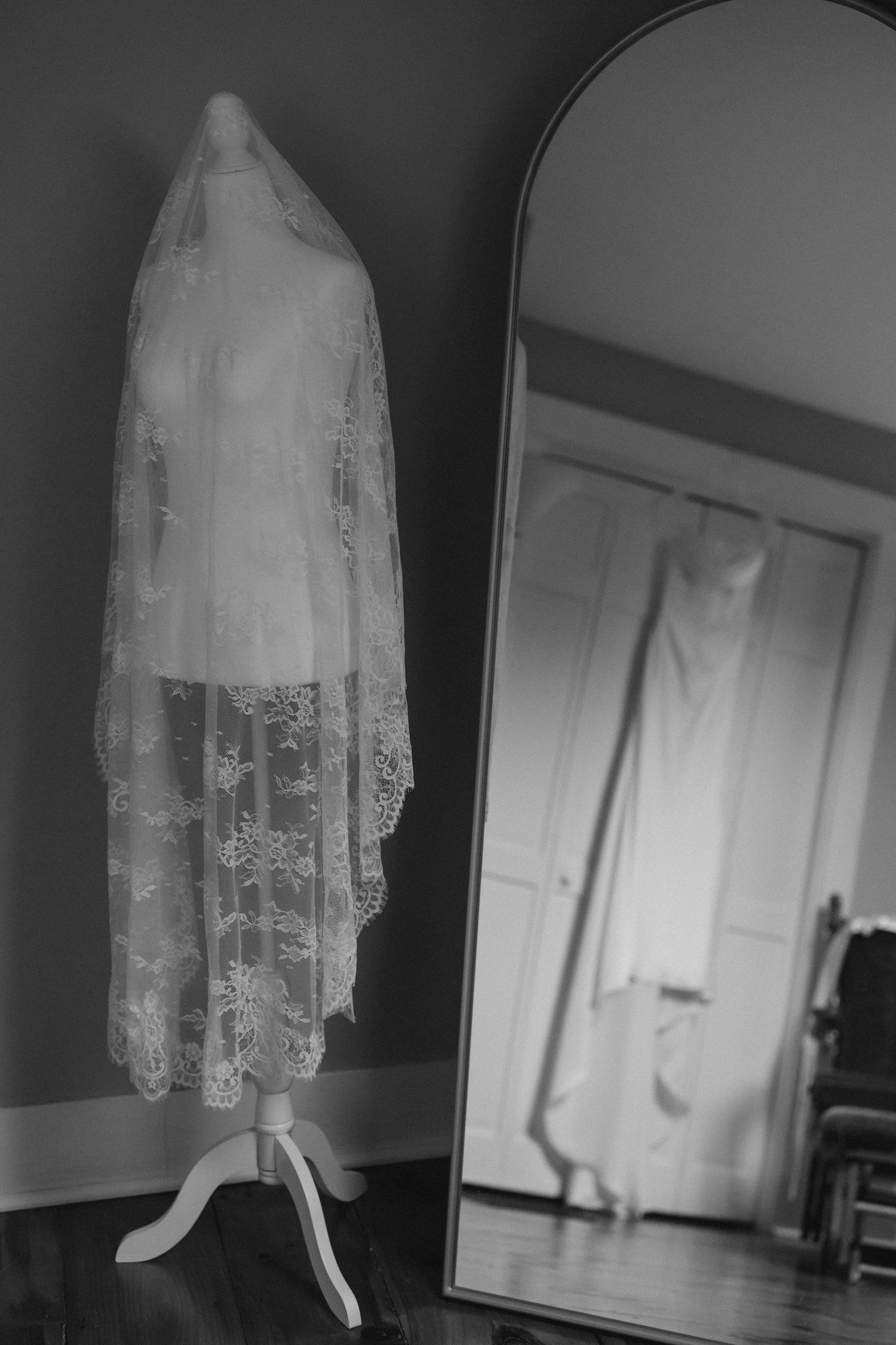 Black and white photo of a lace wedding veil on a mannequin stand beside a tall mirror reflecting a bridal gown.