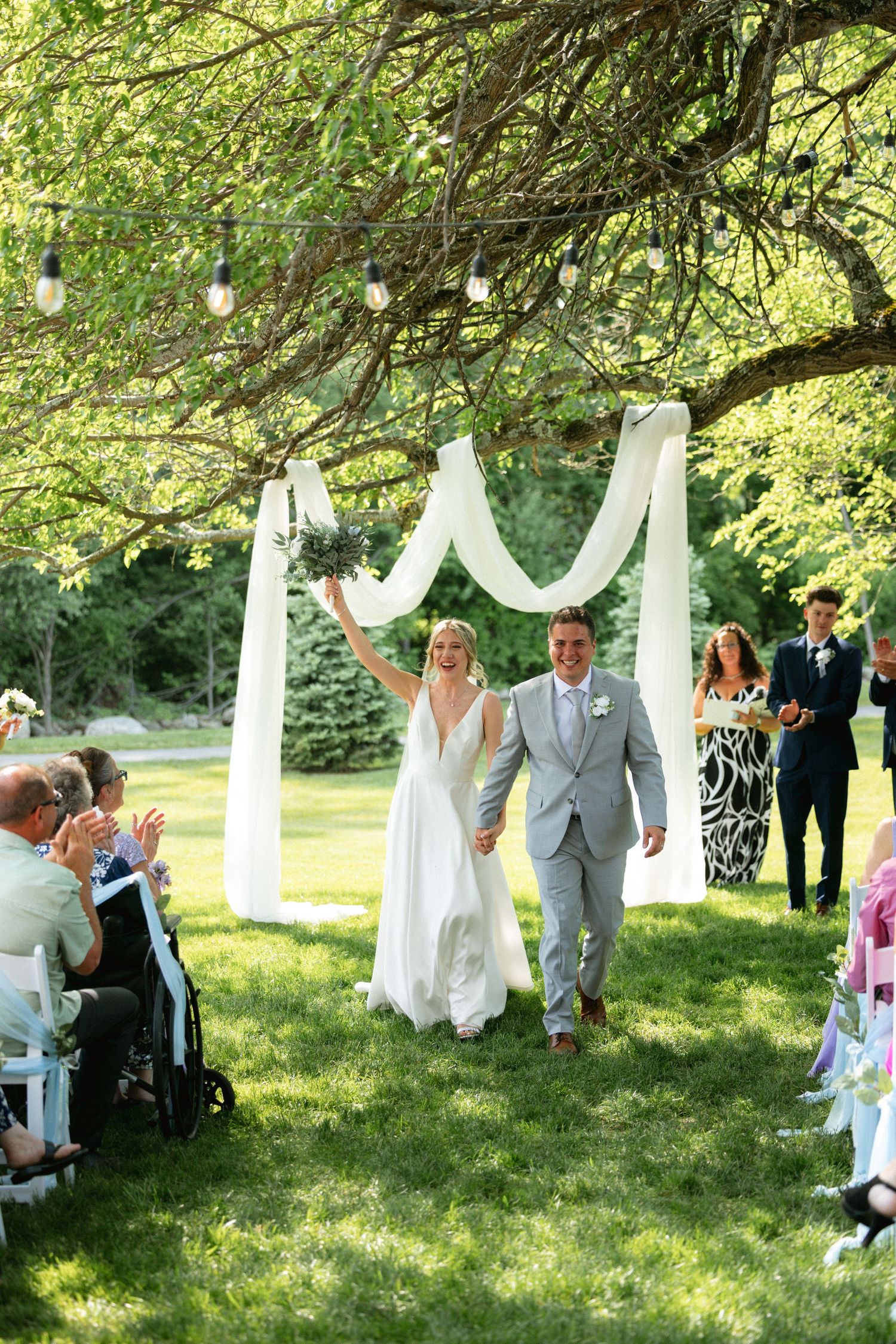 Jubilant bride raises arm as newlyweds walk back down aisle after outdoor wedding ceremony.