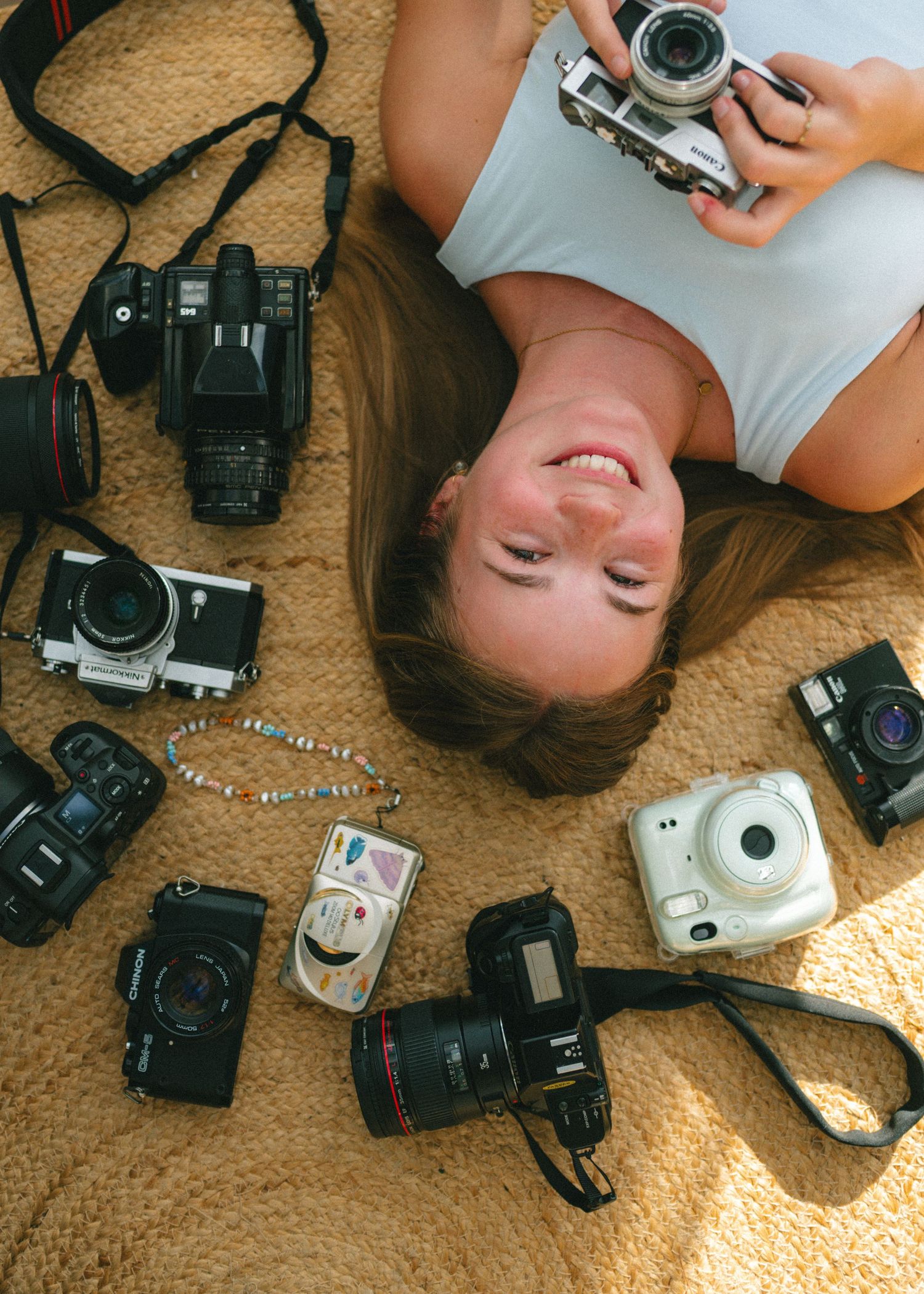 Smiling woman lying on floor surrounded by collection of vintage and modern film cameras and photography equipment.