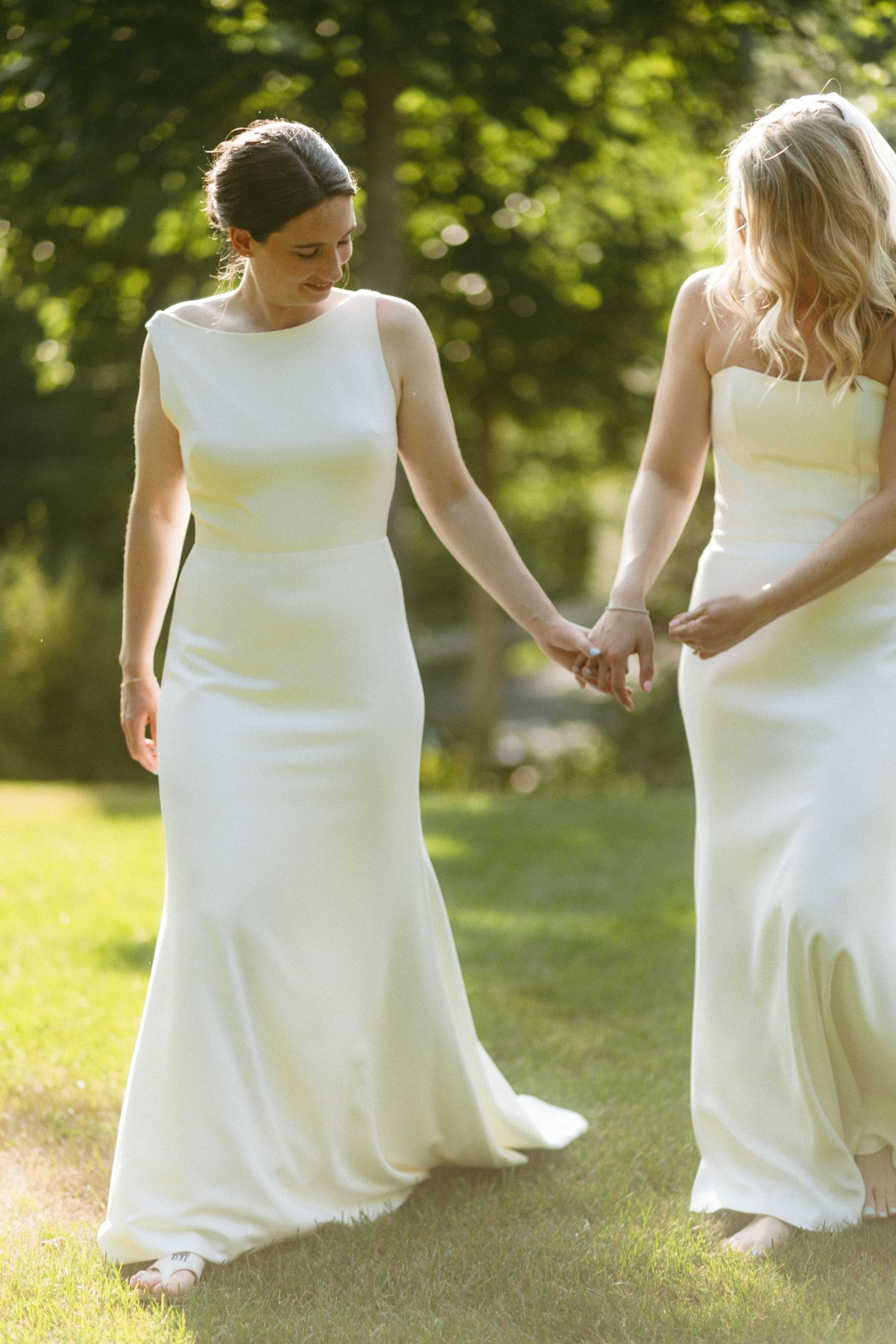 Two people in white dresses hold hands while walking through a sunlit garden setting.