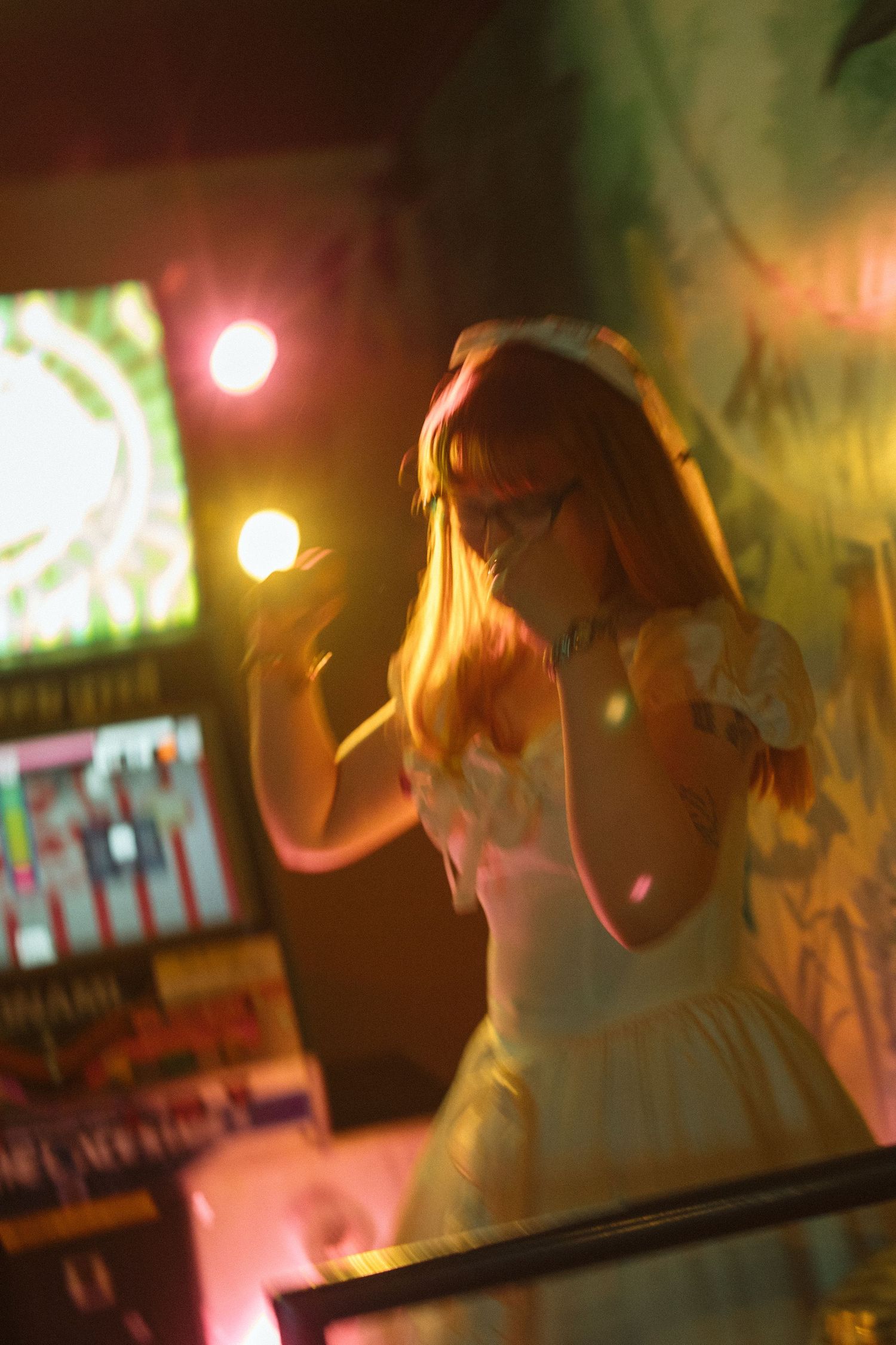 Young woman in white dress holding a glowing orb under colorful neon lights in a dark arcade setting.