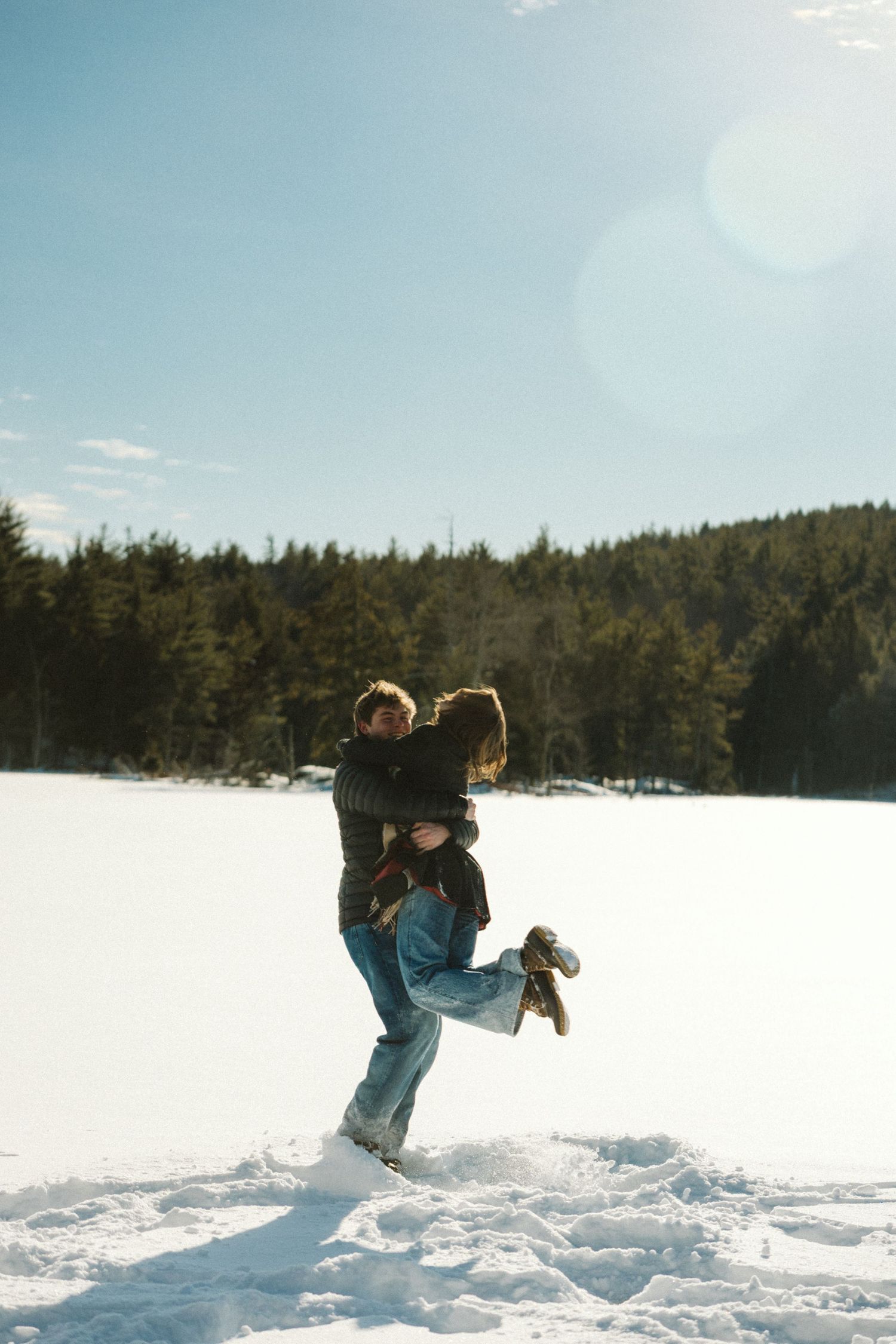 A person in blue winter clothing jumps and plays in deep snow with evergreen trees in the background.