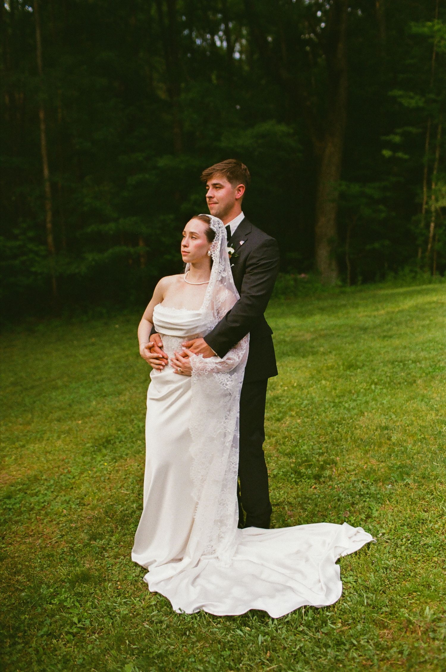 Bride in white gown with long train and groom in black tuxedo posing together on green lawn near forest.