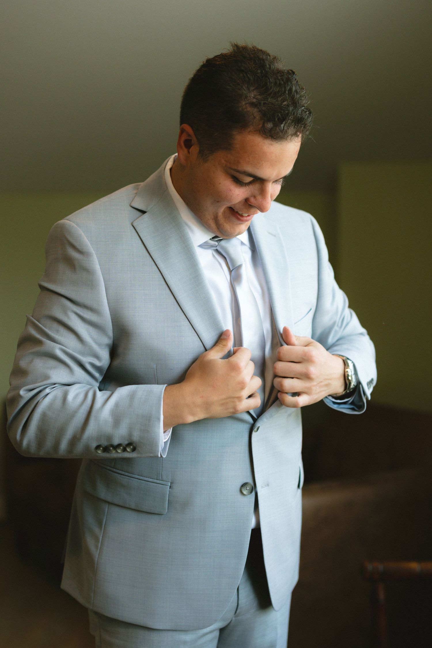A smiling groom in a light gray suit and tie adjusts his jacket lapels while getting ready for his wedding.
