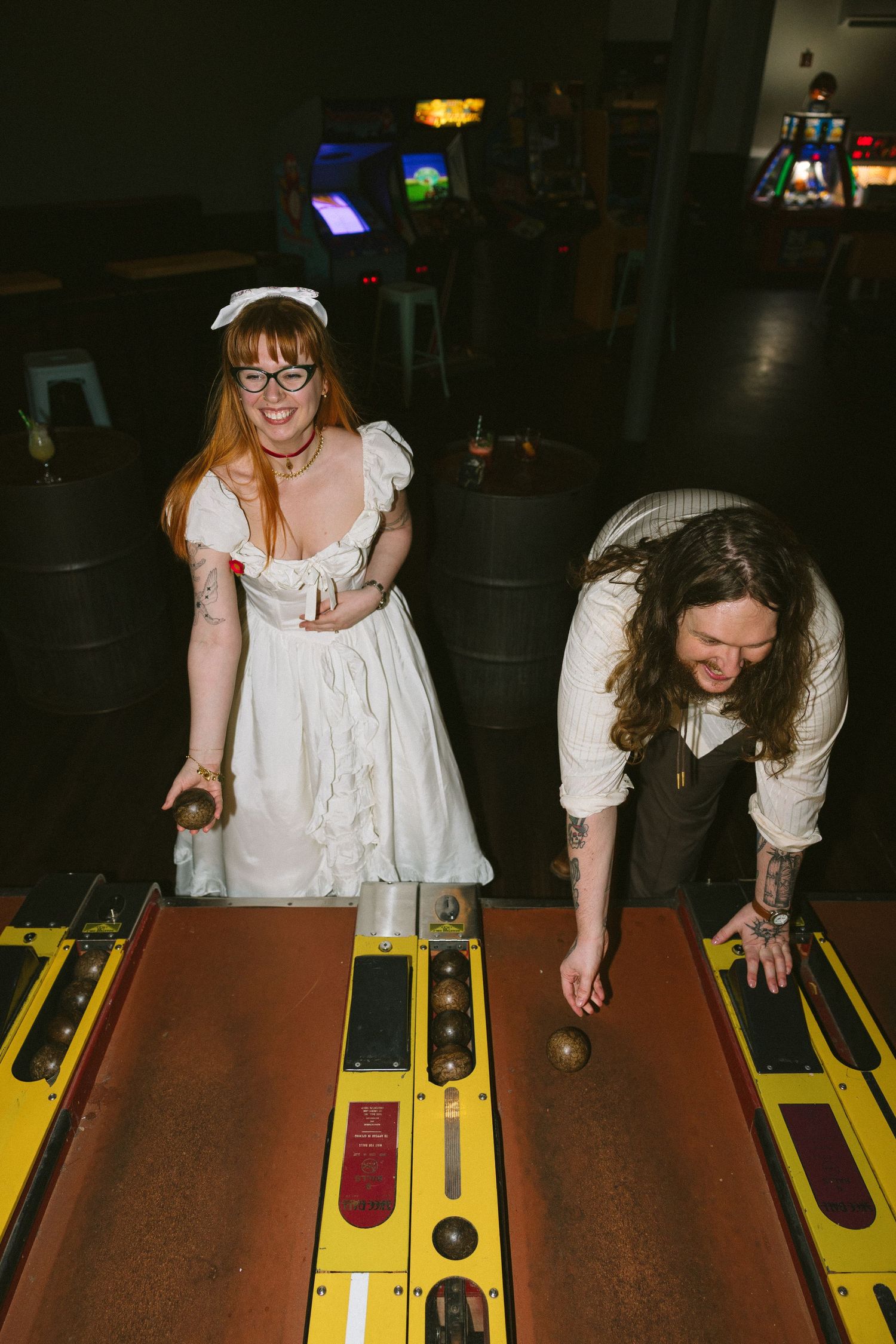 A bride and groom play skee-ball at an arcade wedding reception, surrounded by vintage gaming machines.