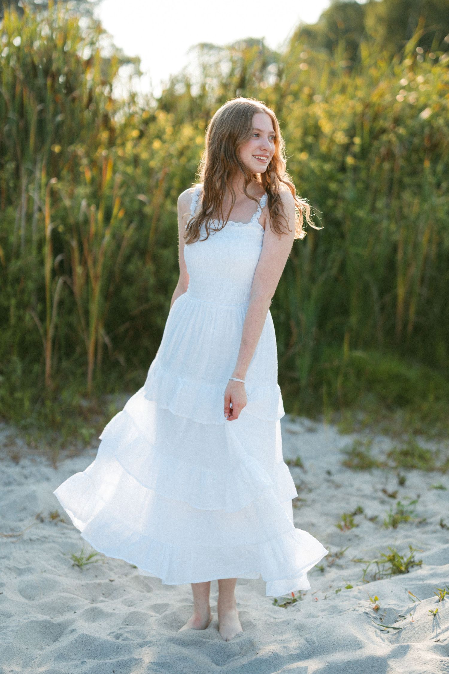 Young person in white dress twirling on sandy beach with tall grass in background.