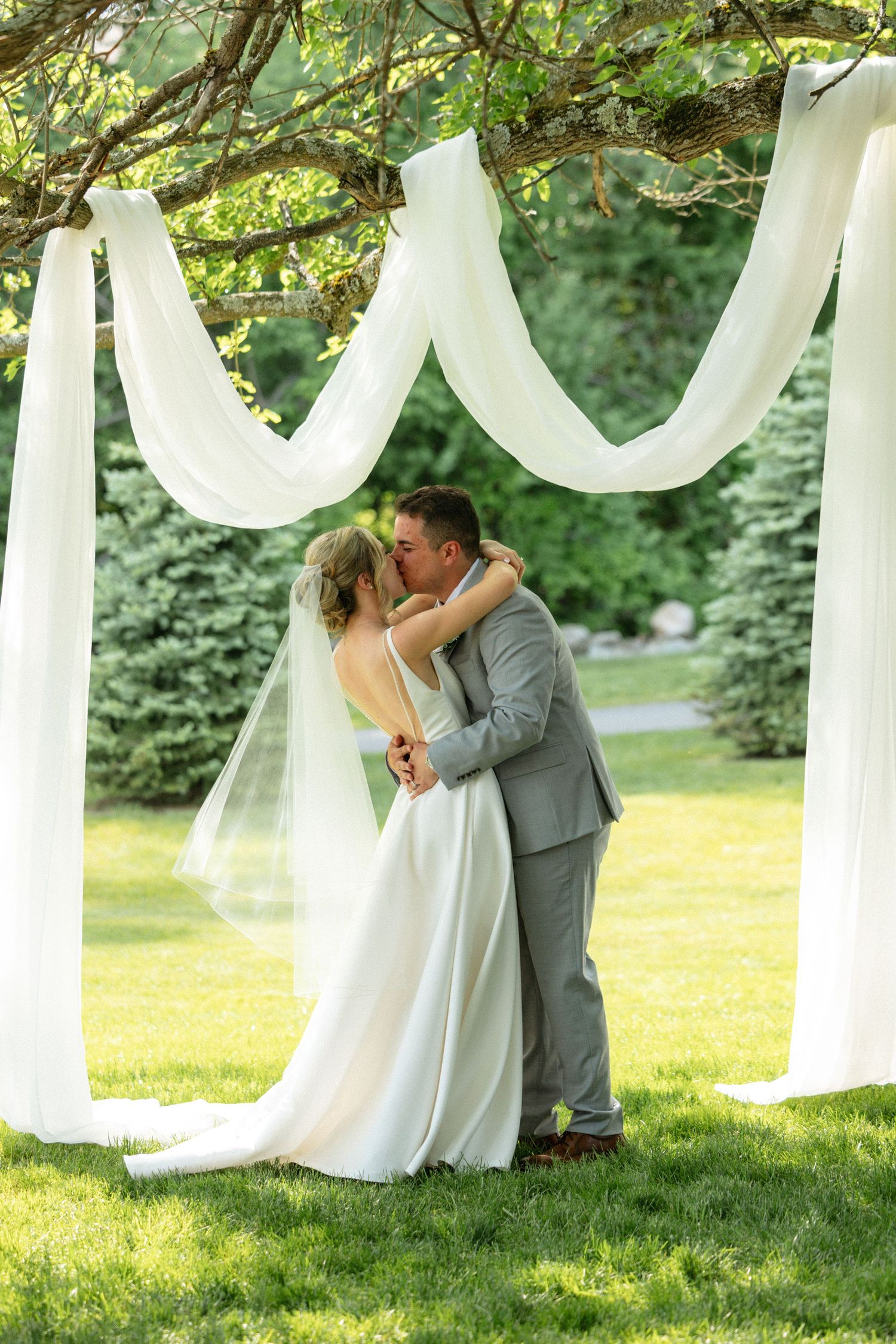 Bride and groom sharing first kiss under white draped fabric outdoor wedding ceremony on lush green lawn.