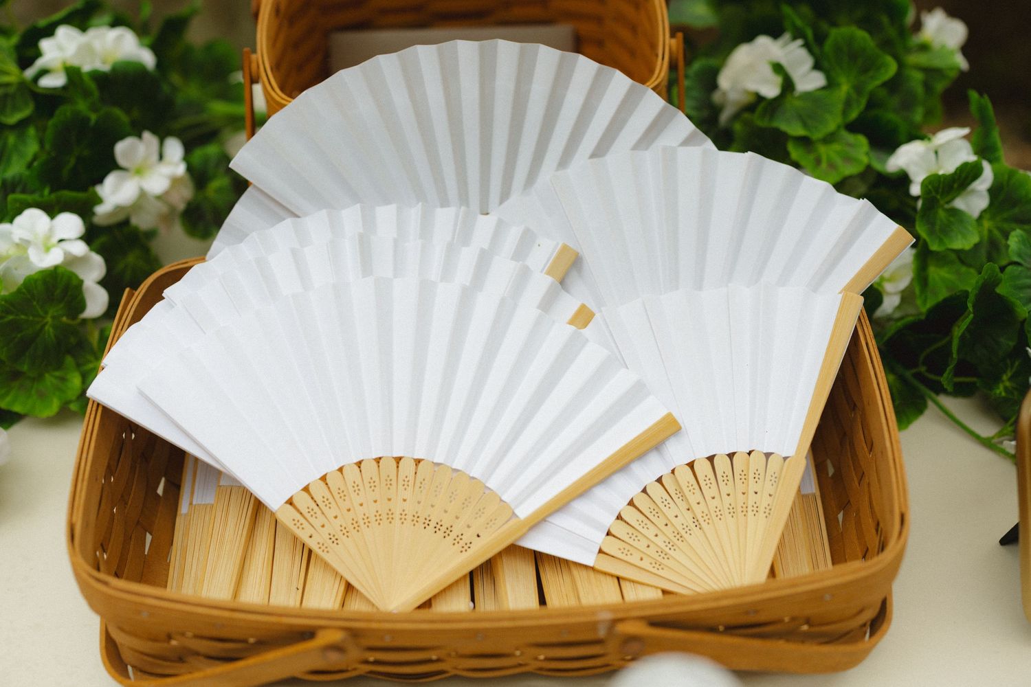 Three white paper hand fans with bamboo handles displayed in a wicker basket with white flowers.