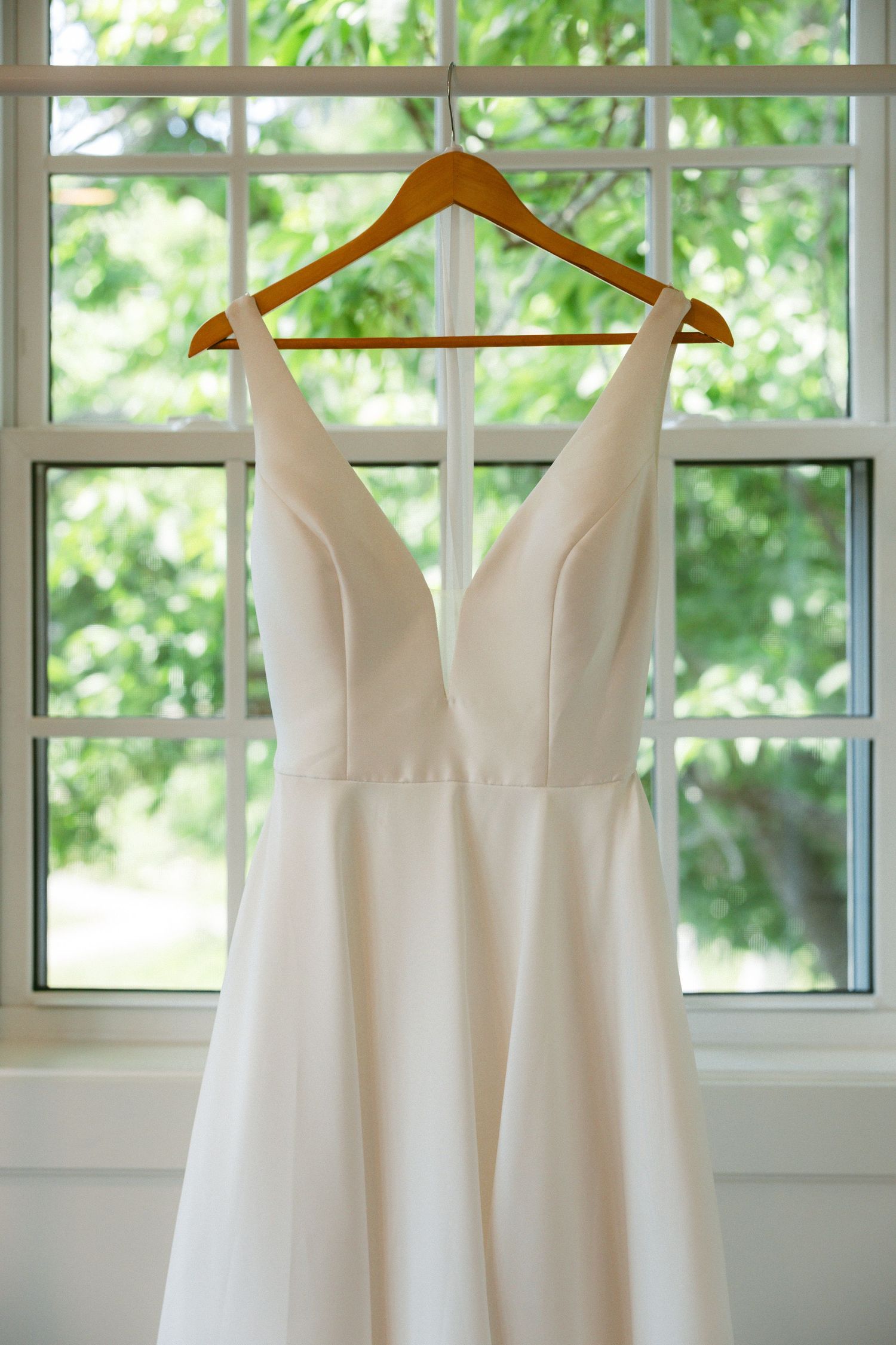White A-line wedding dress with deep V-neckline hanging on wooden hanger in front of bright window with green trees.