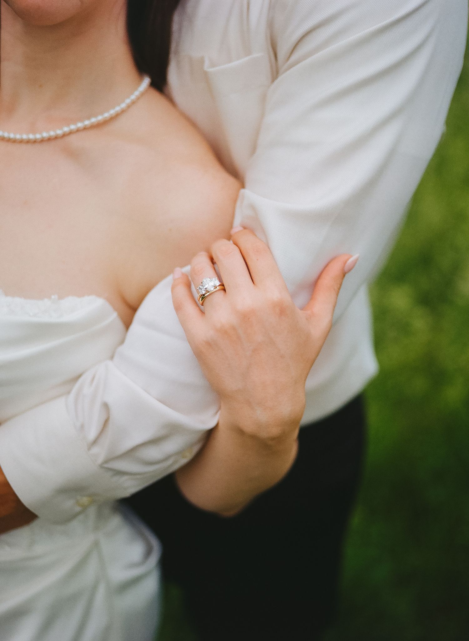 Bride and groom embrace, showcasing a diamond engagement ring and wedding band on her hand, with a pearl necklace visible.