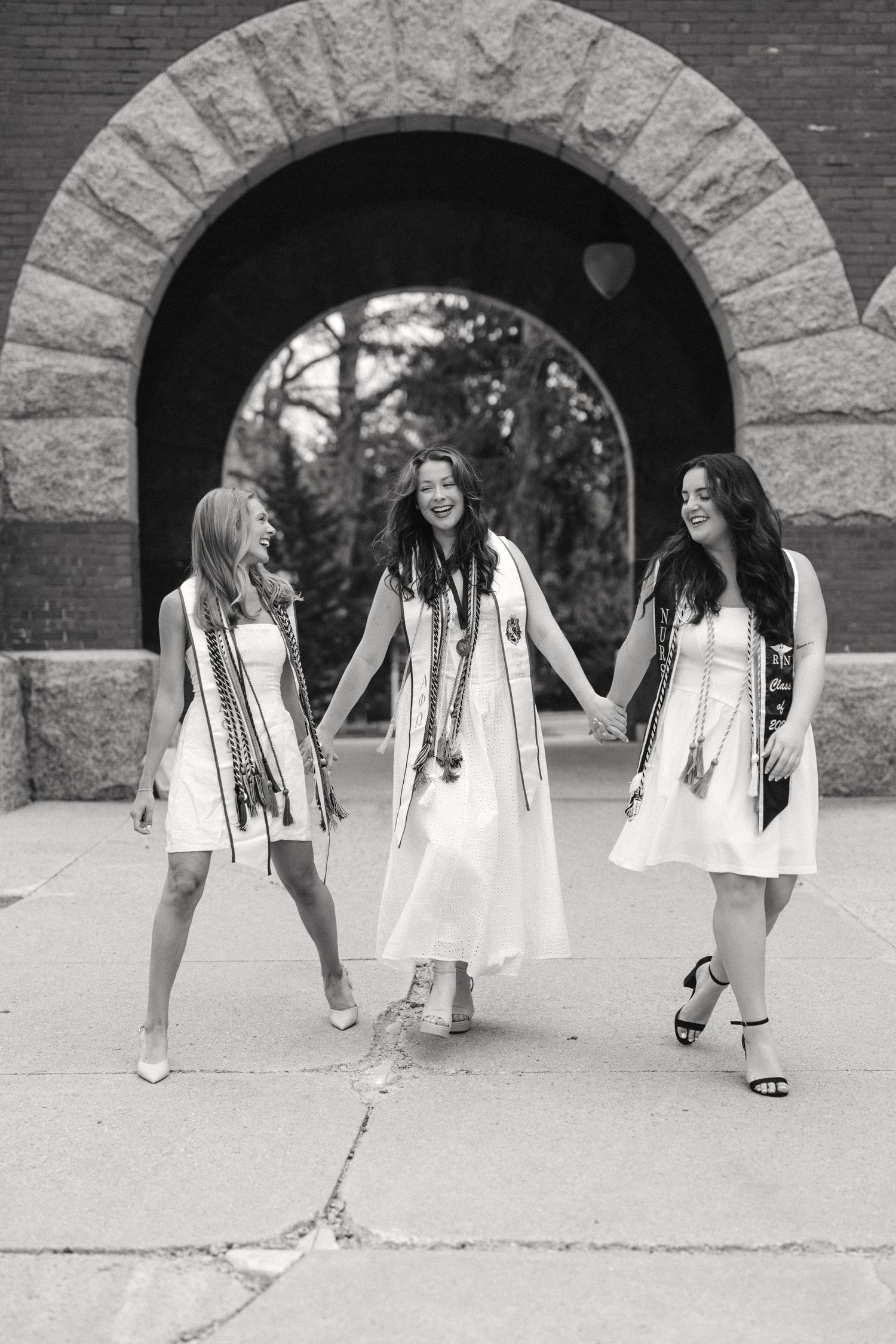 Three women in white graduation dresses and sashes laugh together under a stone arch in a black and white photo.