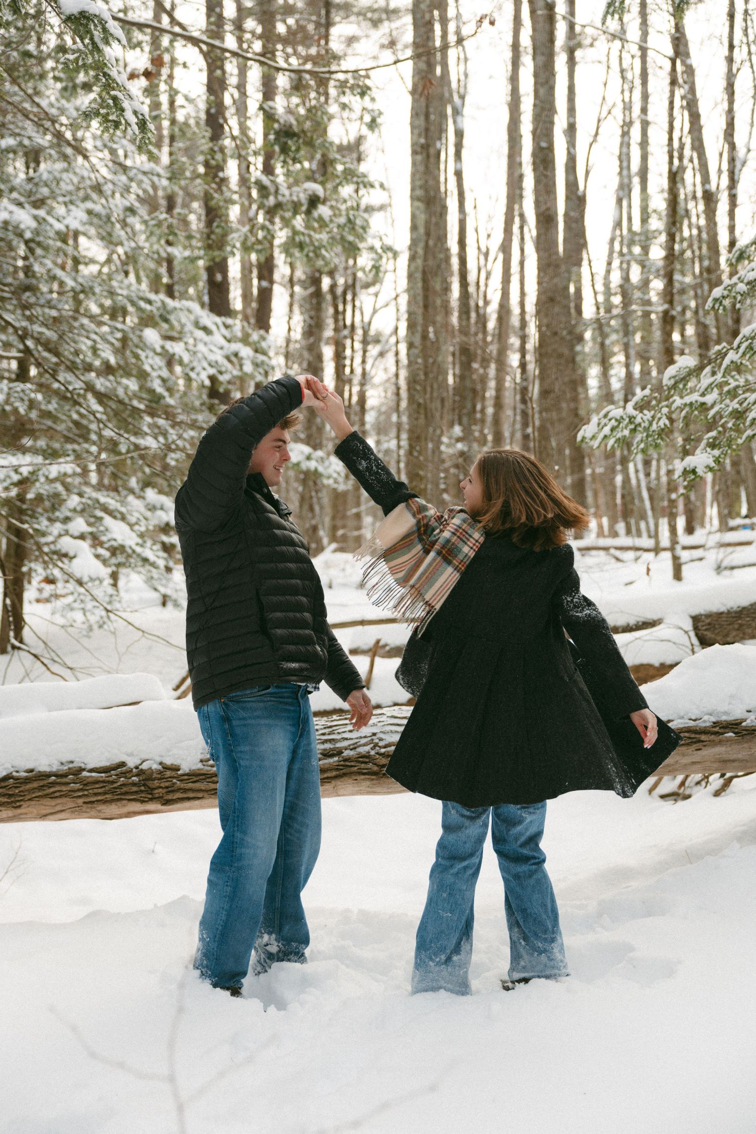 Two people in winter coats dance together in a snowy forest setting with evergreen trees in the background.