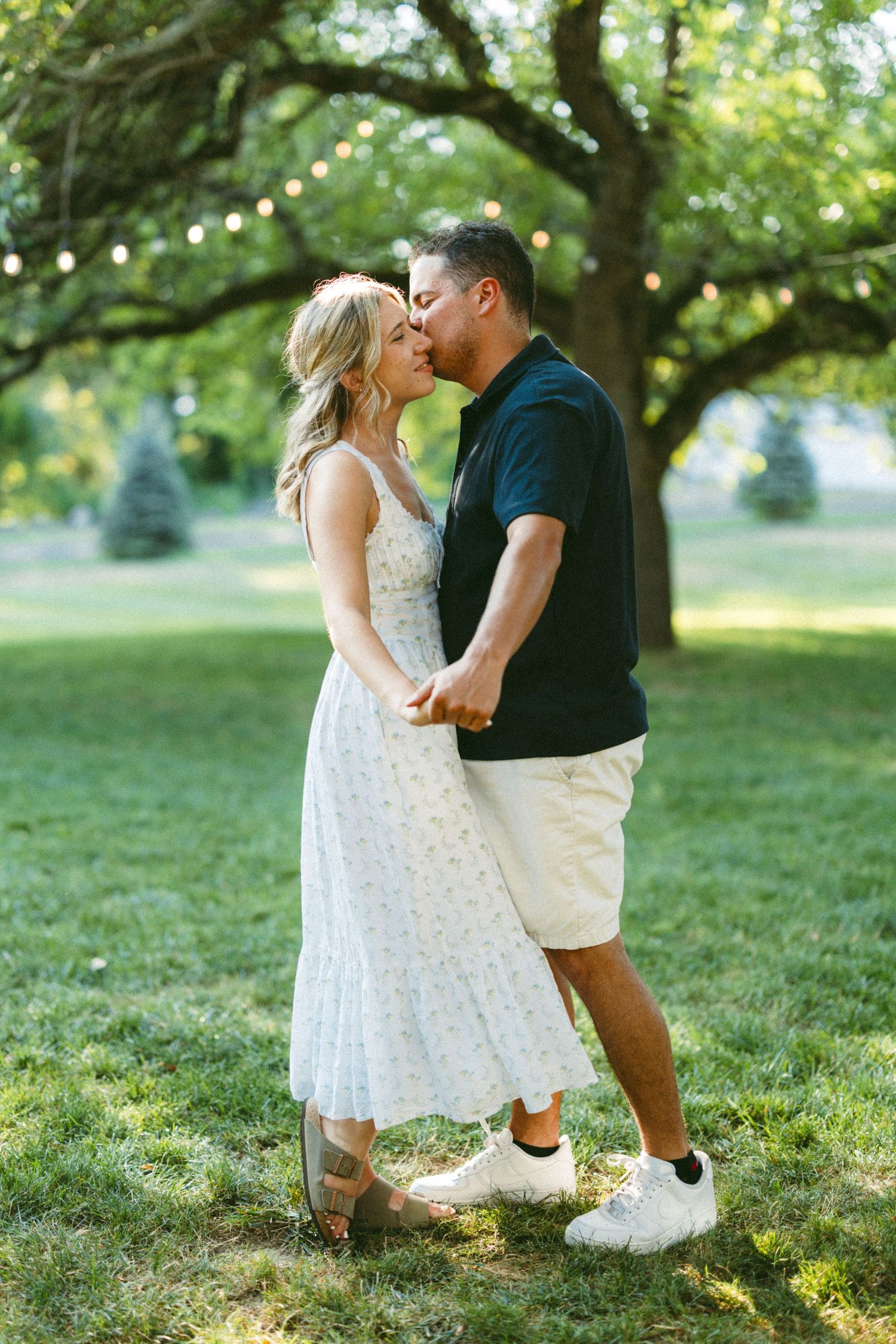 Romantic couple dancing on grass with string lights and trees in the background.