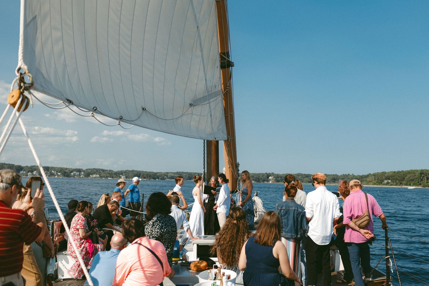 Crowd of wedding guests aboard a sailing vessel with white sails against clear blue skies.