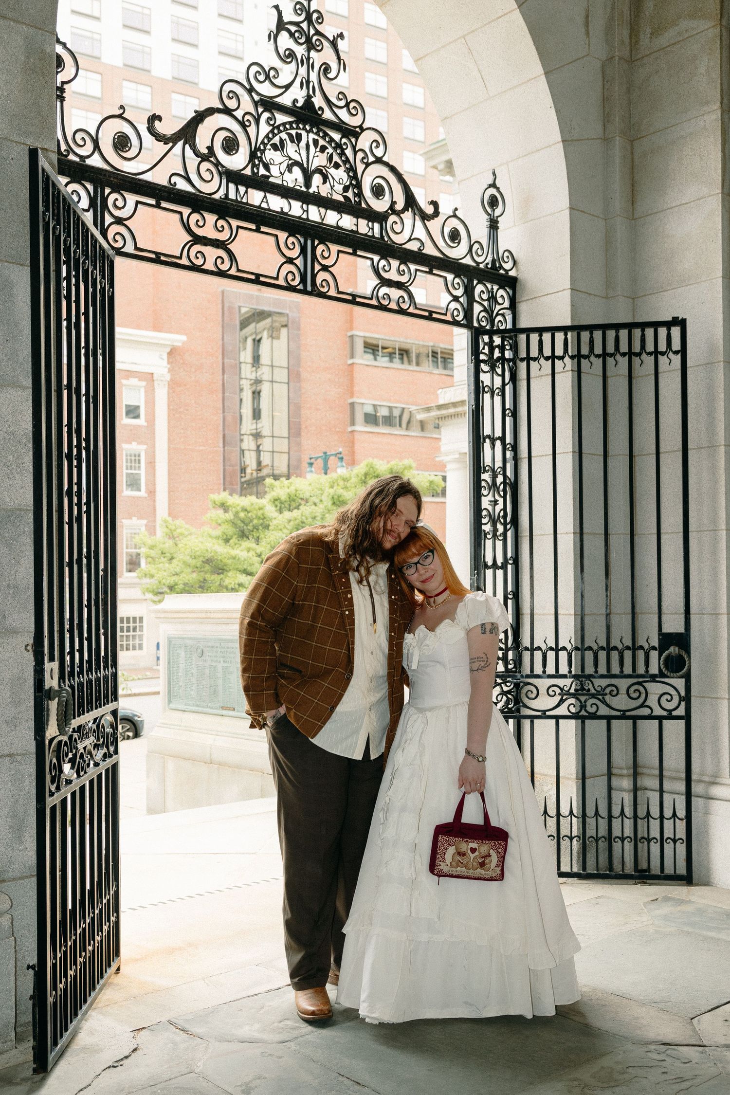 Couple in wedding attire embracing under ornate iron gates at a historic stone building.