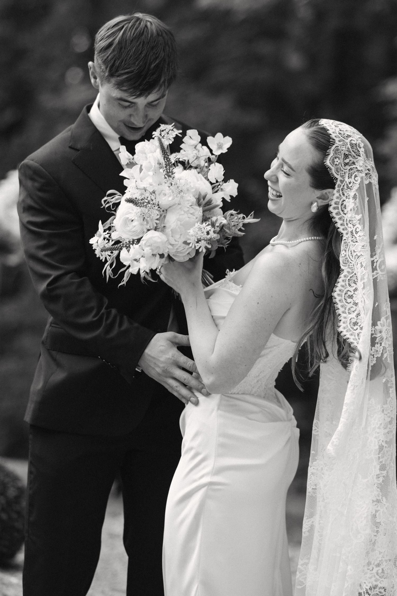 Black and white photo of bride and groom sharing joyful moment with floral bouquet outdoors.
