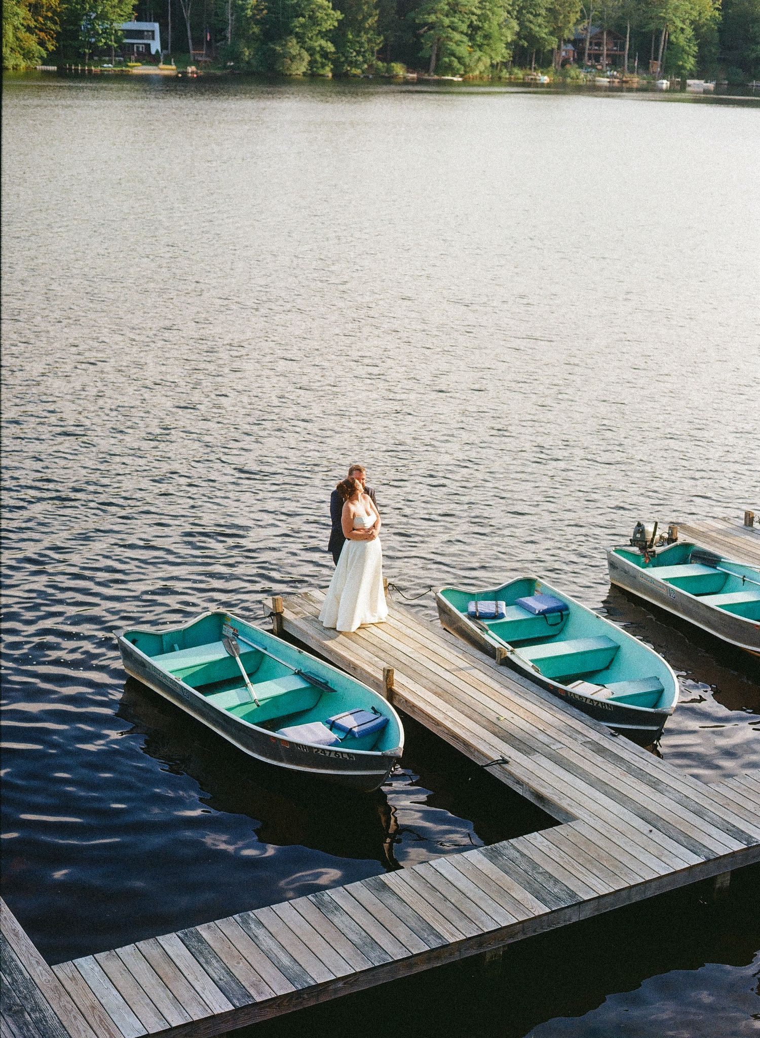 Bride and groom embrace on a wooden dock surrounded by turquoise rowboats on a calm lake.