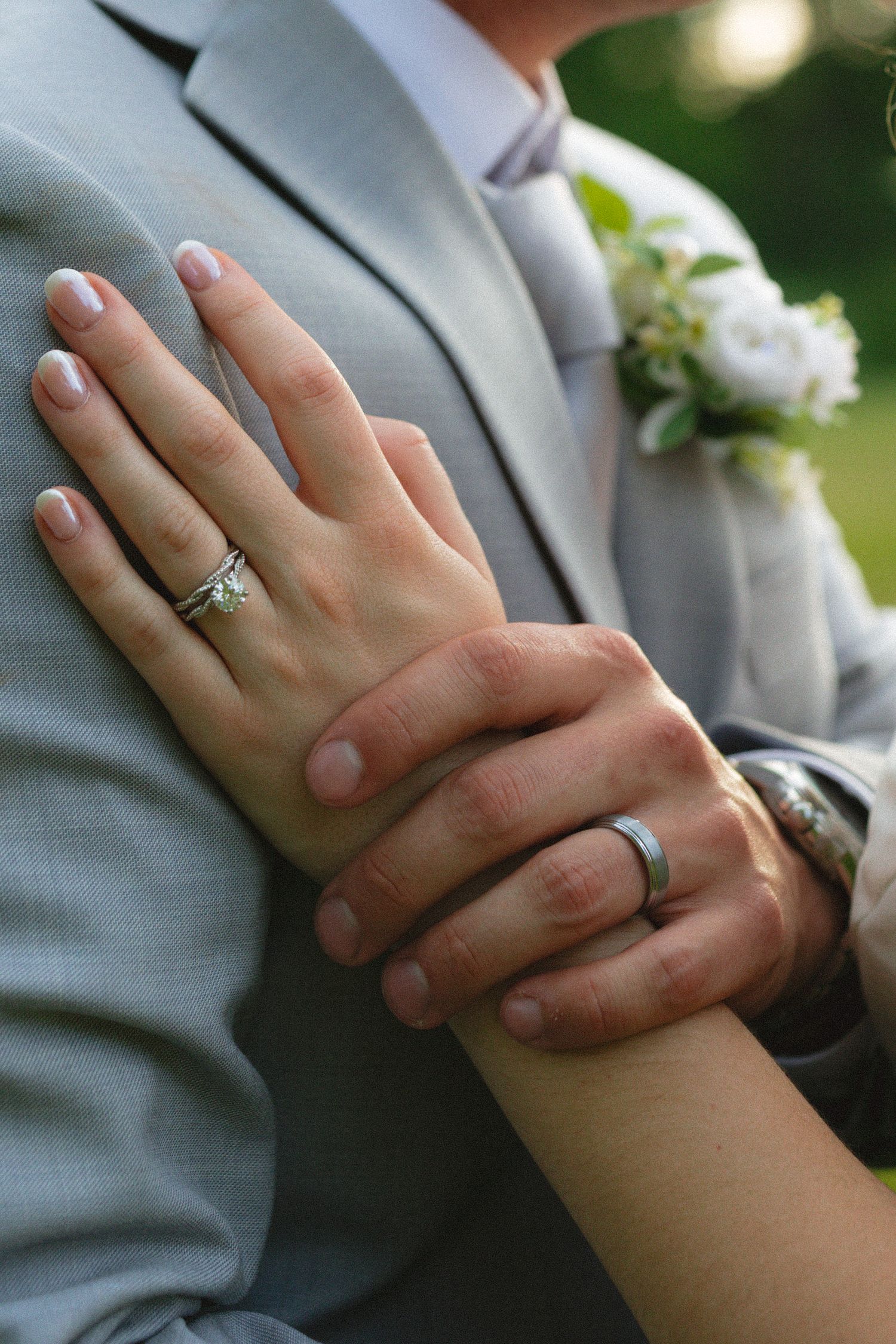Close-up of newlywed couple's hands showing wedding rings, with groom in gray suit and boutonniere.