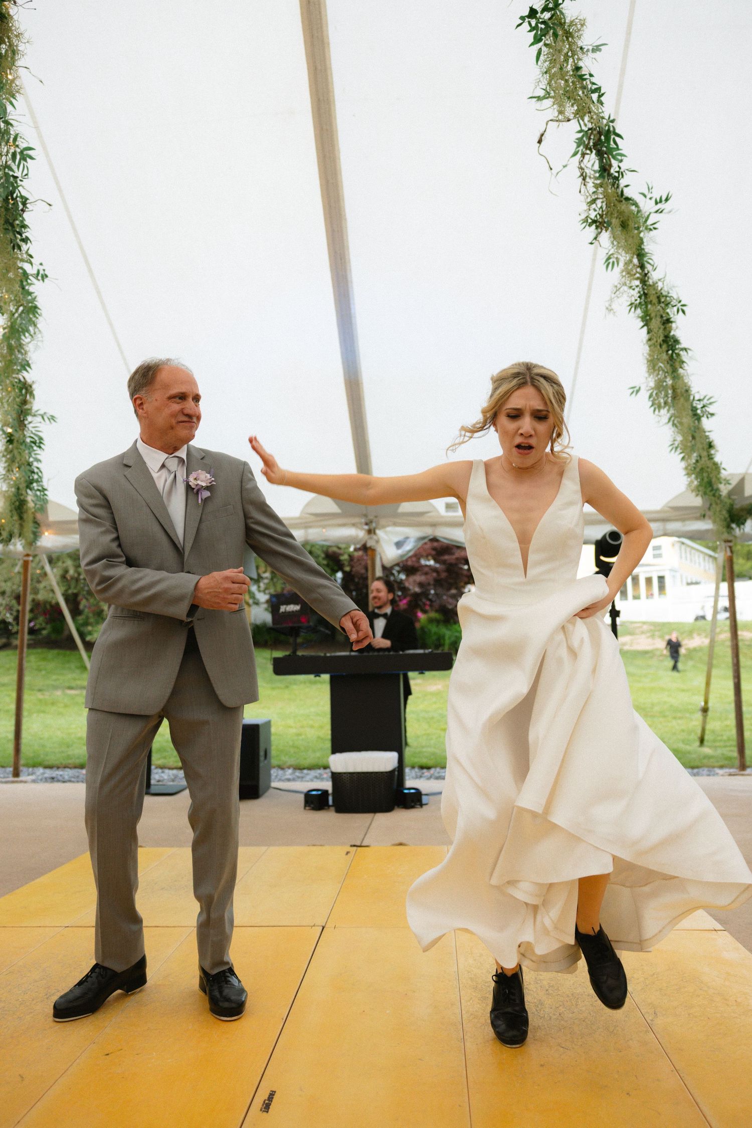 Bride in white gown dances energetically with older man in gray suit under decorated tent at wedding reception.