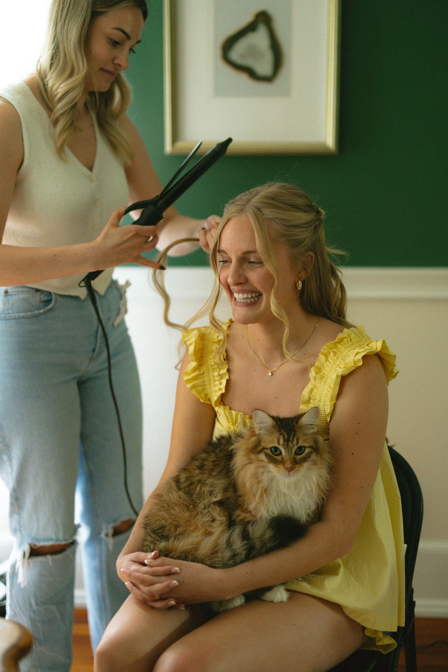 Woman in yellow dress holds fluffy cat while another woman styles her hair with a curling iron indoors.