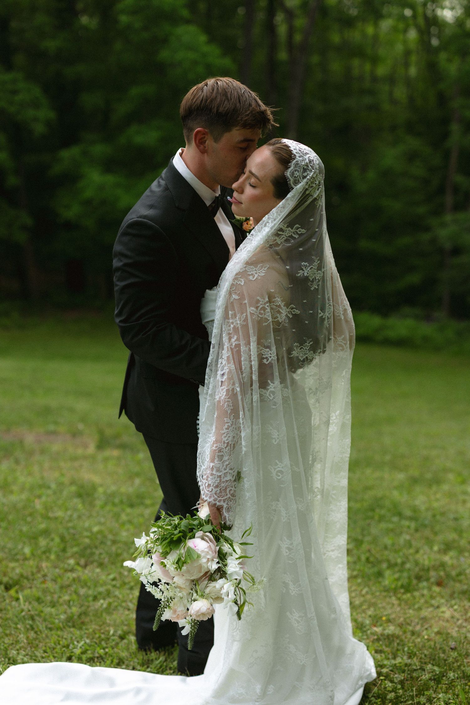 Groom kisses bride on forehead as she holds a white floral bouquet in a lush green outdoor setting.