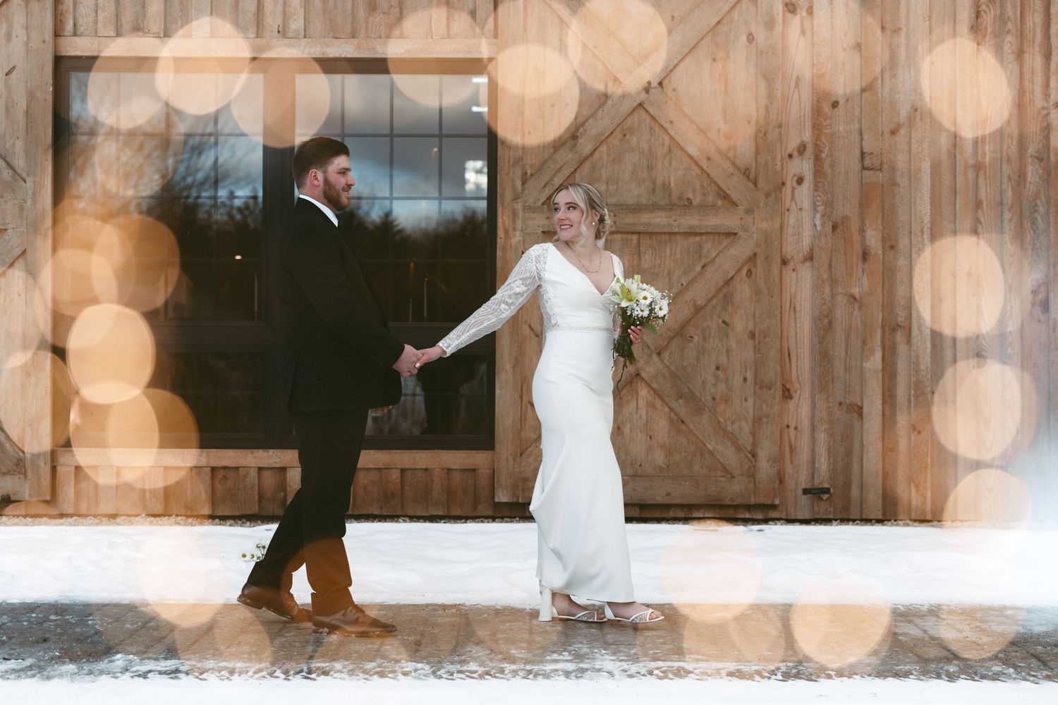 Newlywed couple dances joyfully on rustic wooden dance floor with romantic bokeh lights glowing in background.