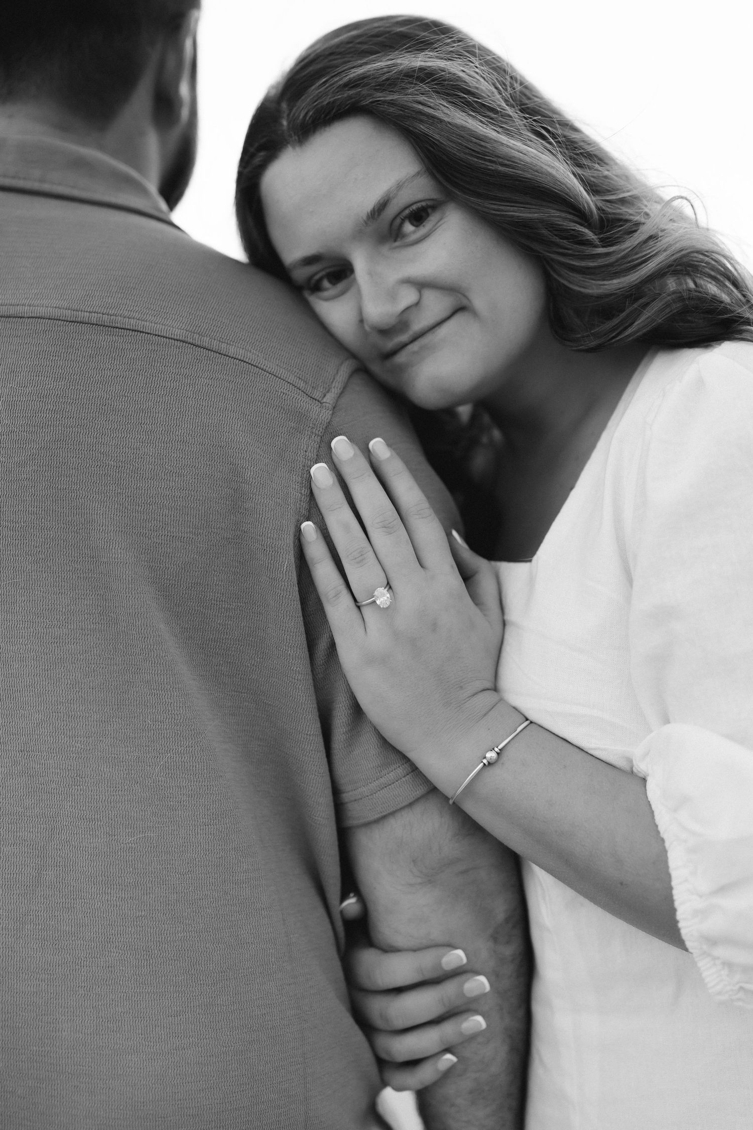Black and white photo of a smiling woman resting her head on a mans shoulder, showing off her engagement ring.