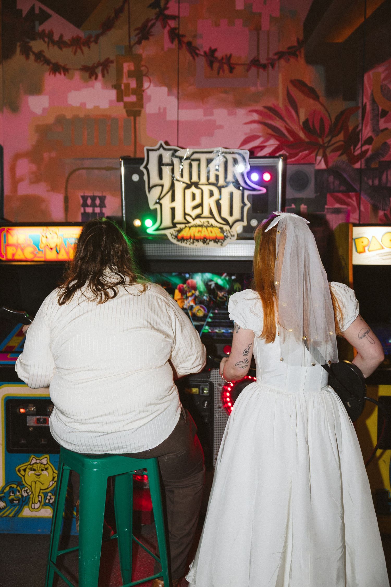 Bride in white dress and veil plays Guitar Hero arcade game with friend at wedding reception.