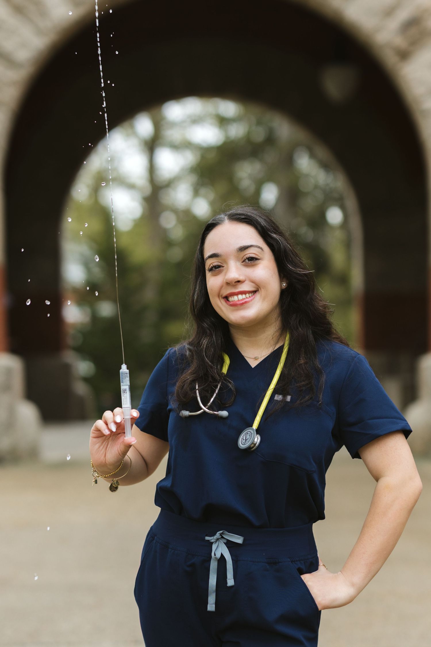 Smiling nurse in navy scrubs holding a syringe outdoors with a stethoscope around her neck.