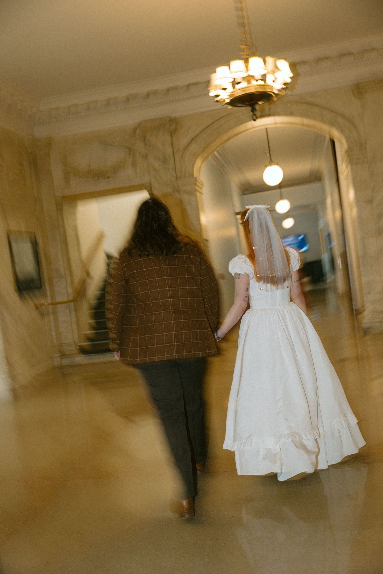 Bride in white gown and veil walks with guest through elegant marble hallway with chandelier.