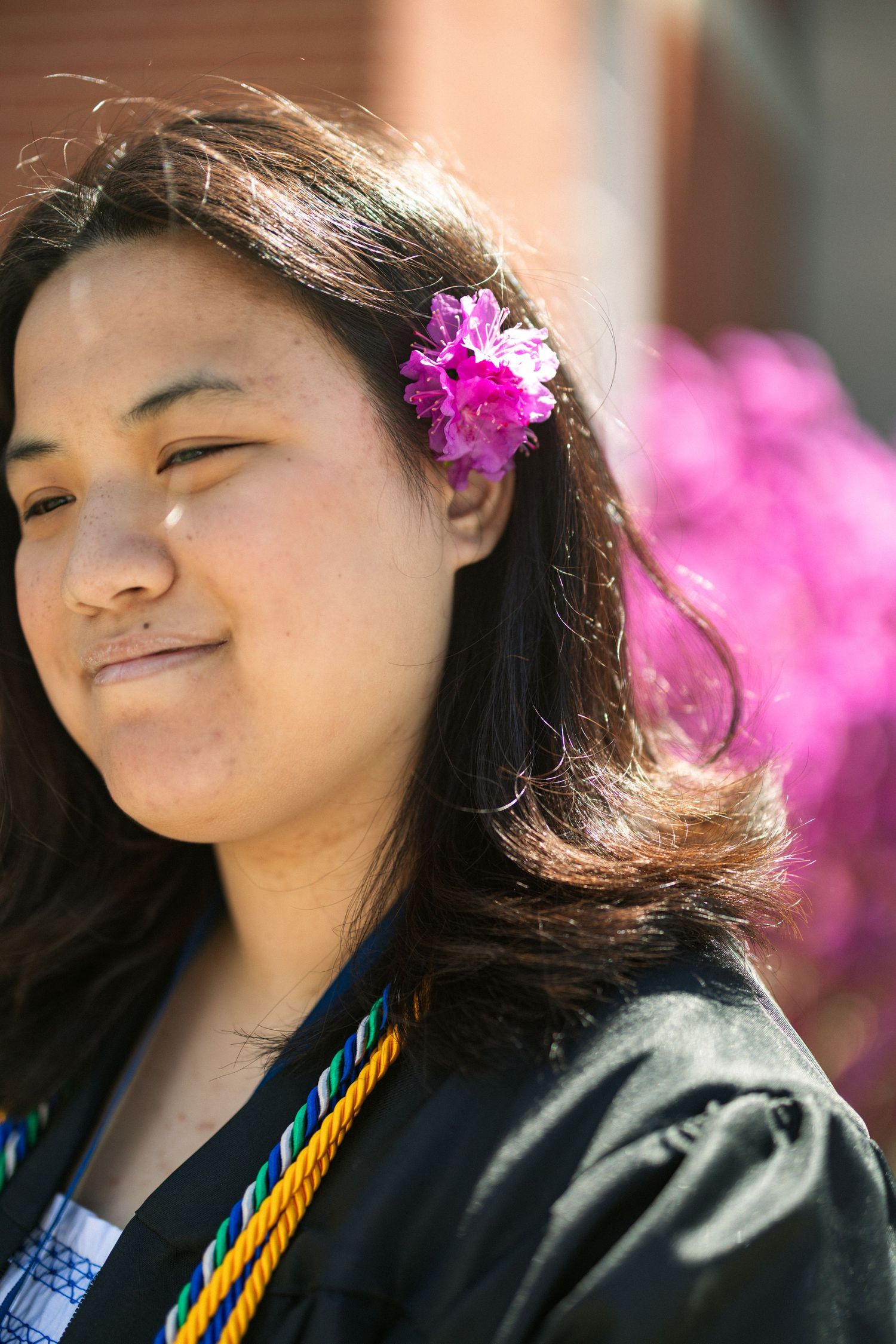 Young woman in graduation gown with pink flower in hair, smiling outdoors near blooming flowers.