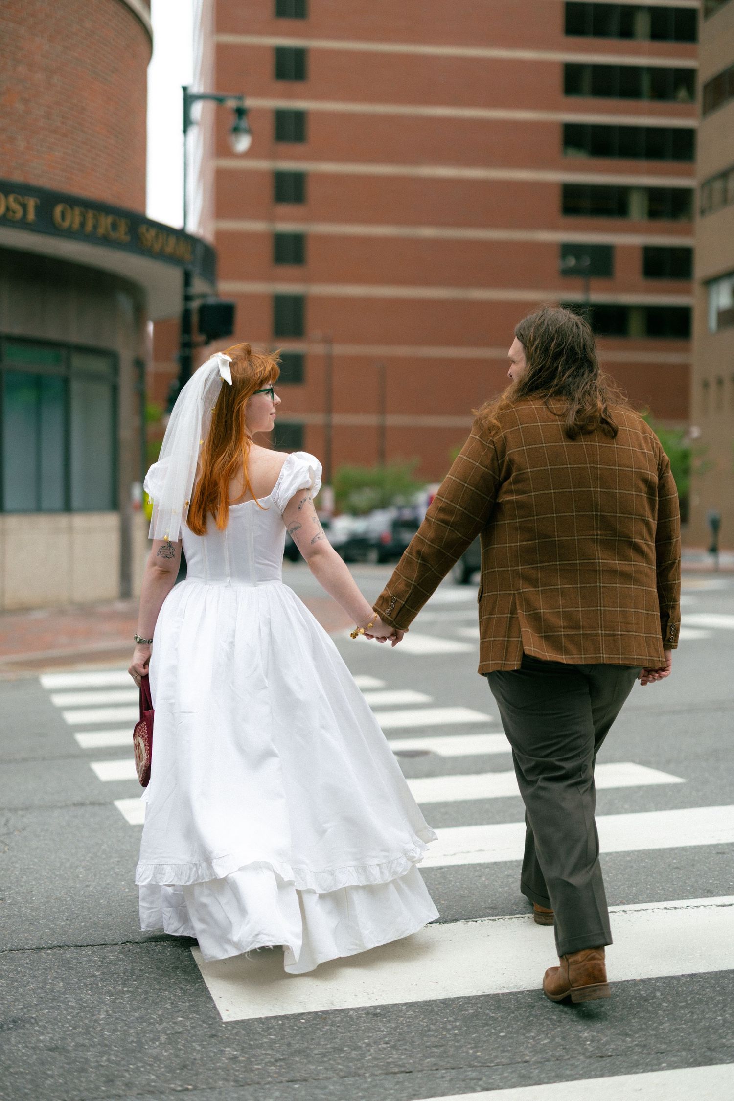 A bride in white dress and groom in brown jacket hold hands crossing a zebra crossing in an urban street setting.