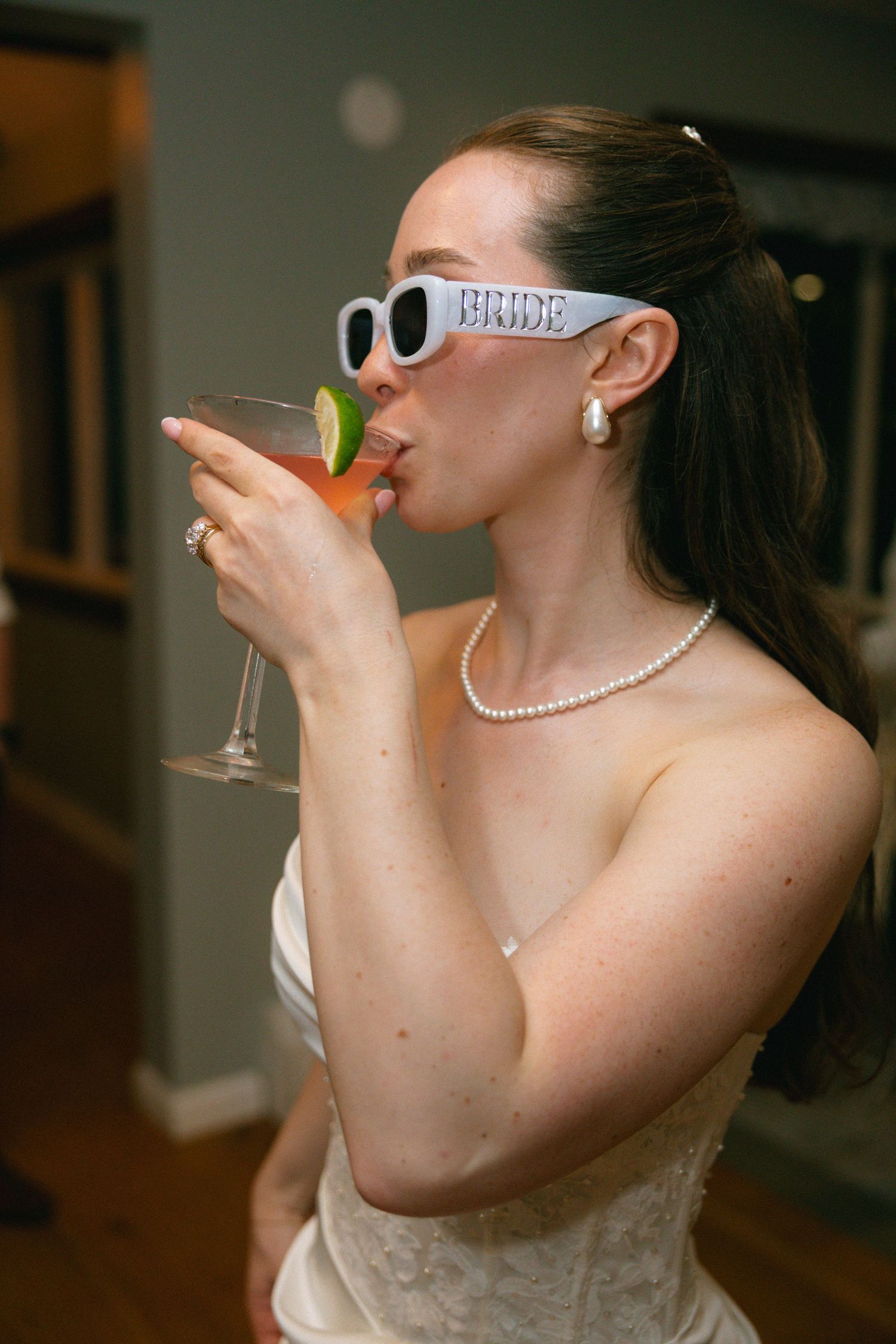 Bride wearing white sunglasses and pearl jewelry sipping a cocktail garnished with lime at her wedding reception.