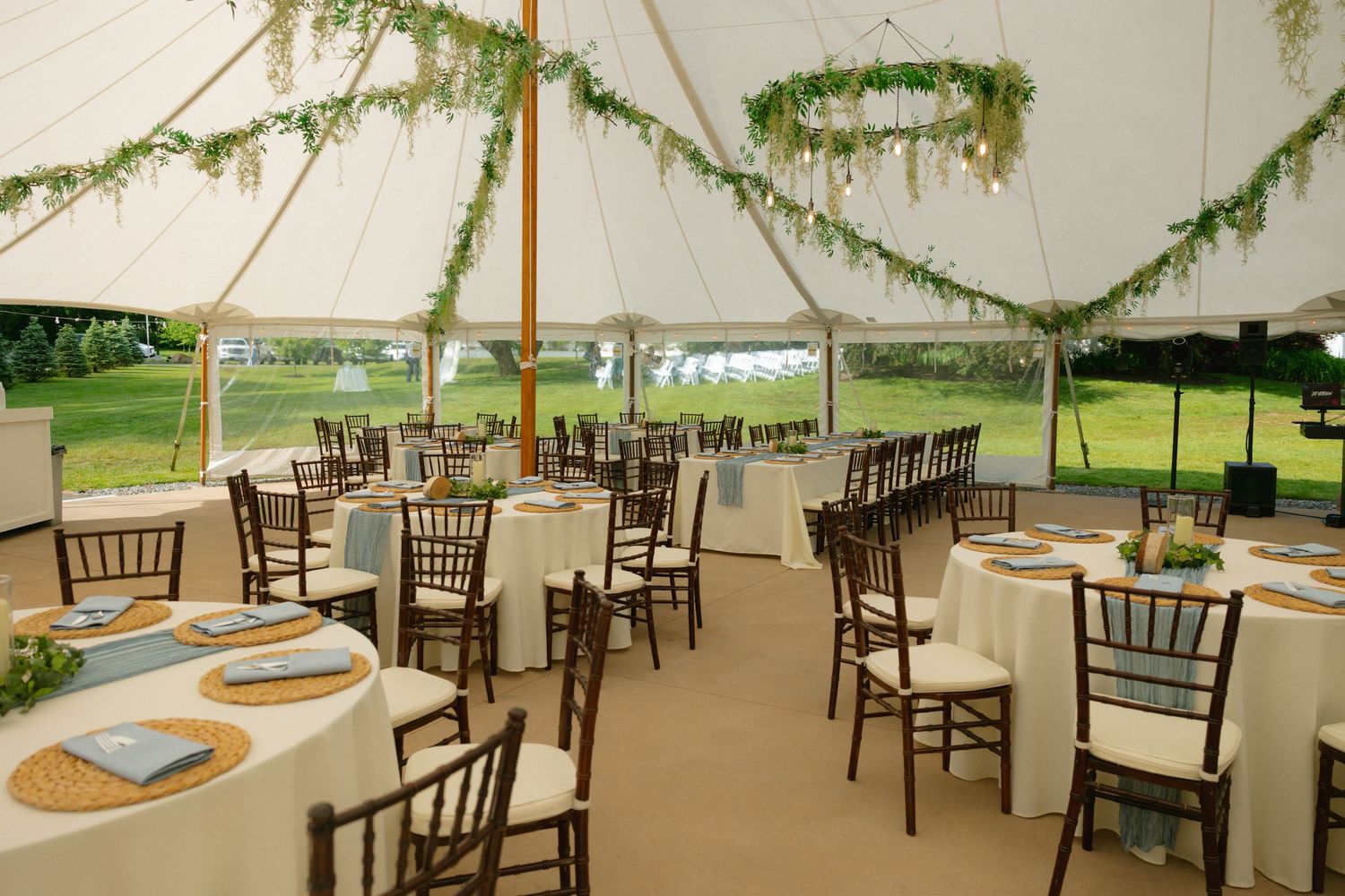 Elegant outdoor wedding tent with white linens, chiavari chairs, and lush greenery garlands draped from the ceiling.