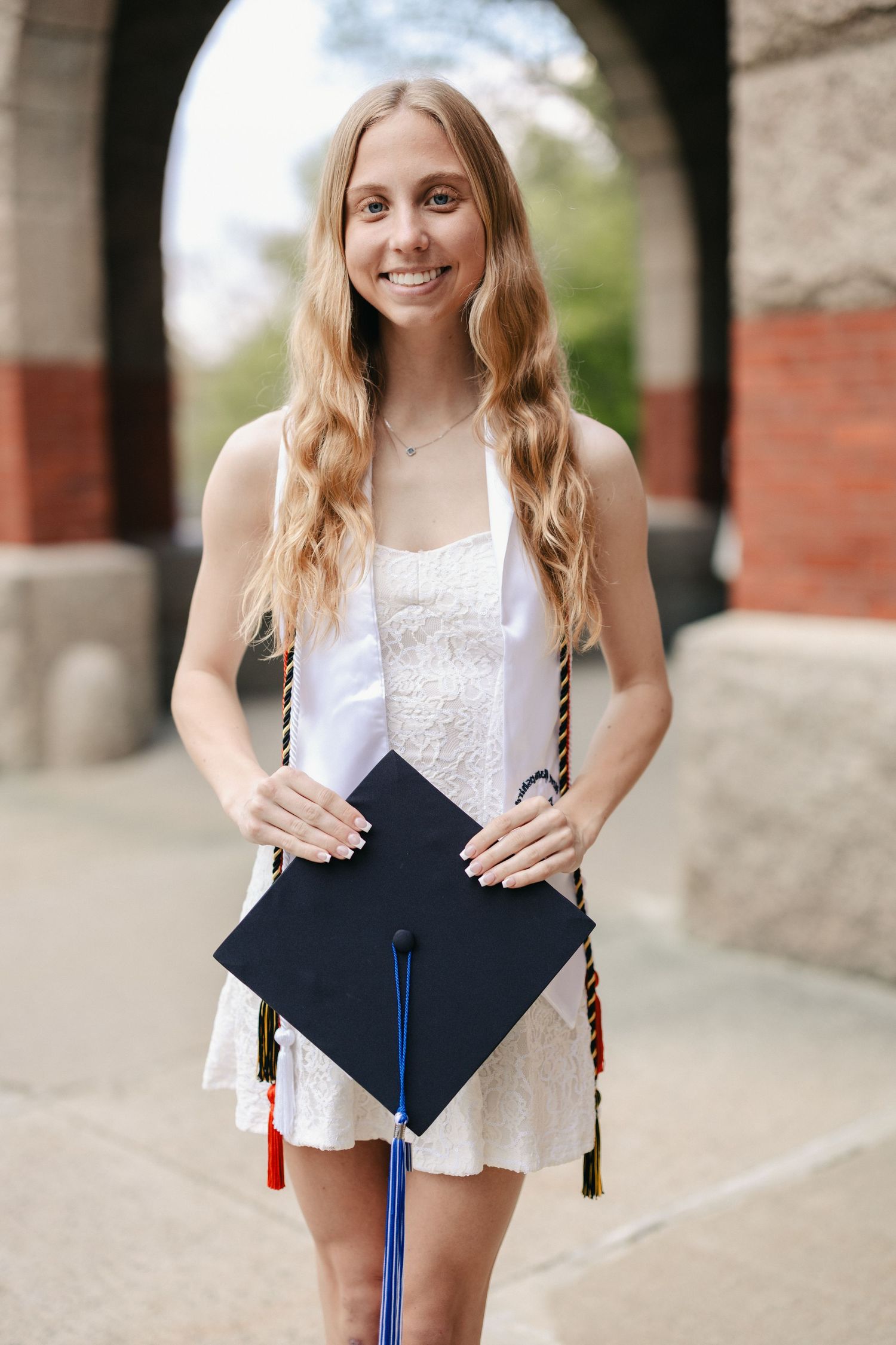 Graduate in lace white dress poses with decorated cap under stone archway.
