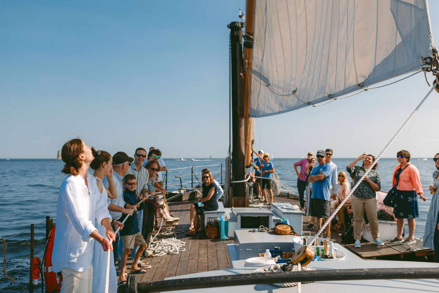 A group gathers on a sailboat deck during a sunny day on the water with white sails visible against blue sky.