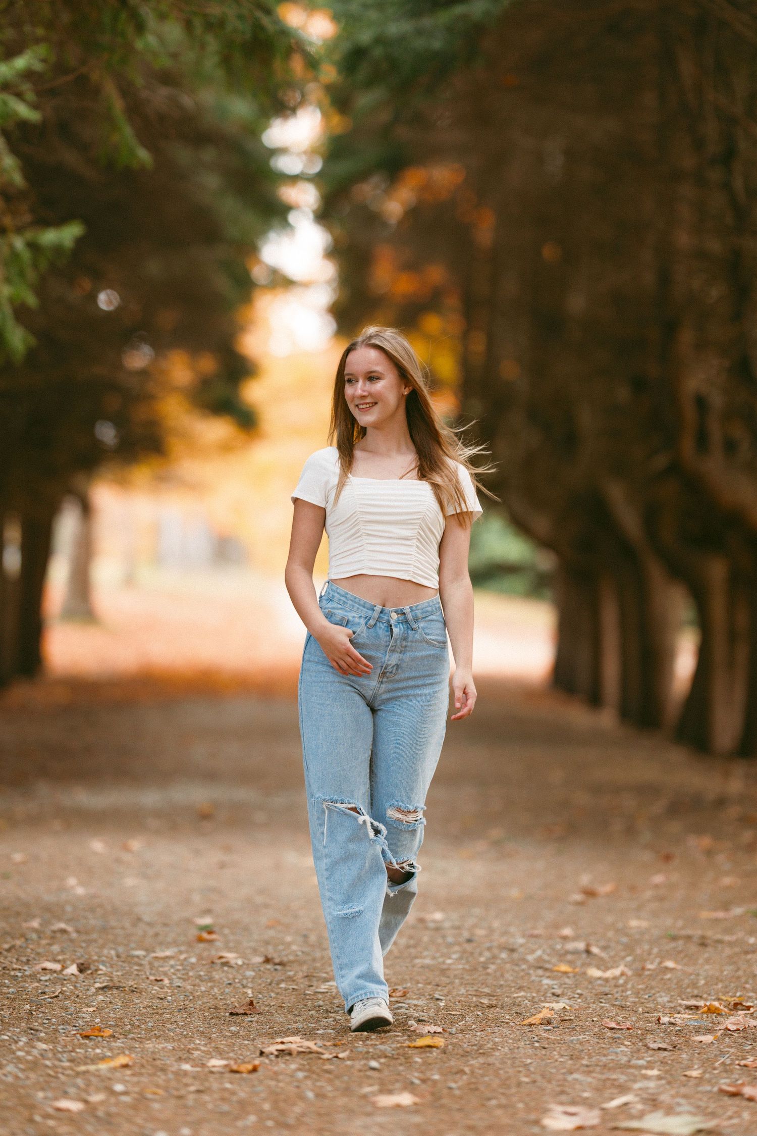 Senopr walking on tree-lined path wearing ripped jeans and white top in golden sunlight.